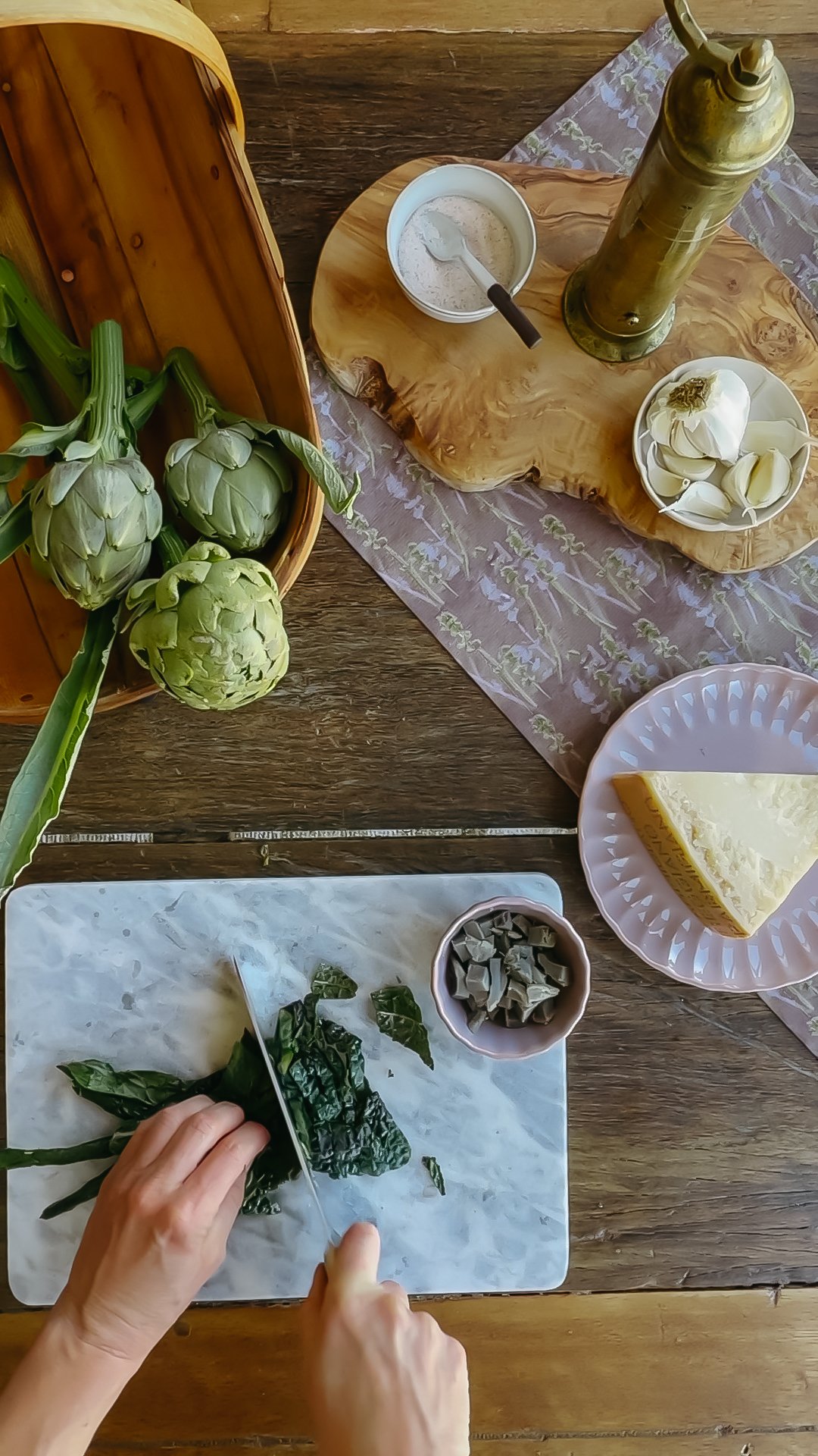 Person chopping green herbs on a white cutting board with artichokes and vegetables in the background. Various ingredients and kitchen tools are arranged on a wooden table and a decorative cloth.