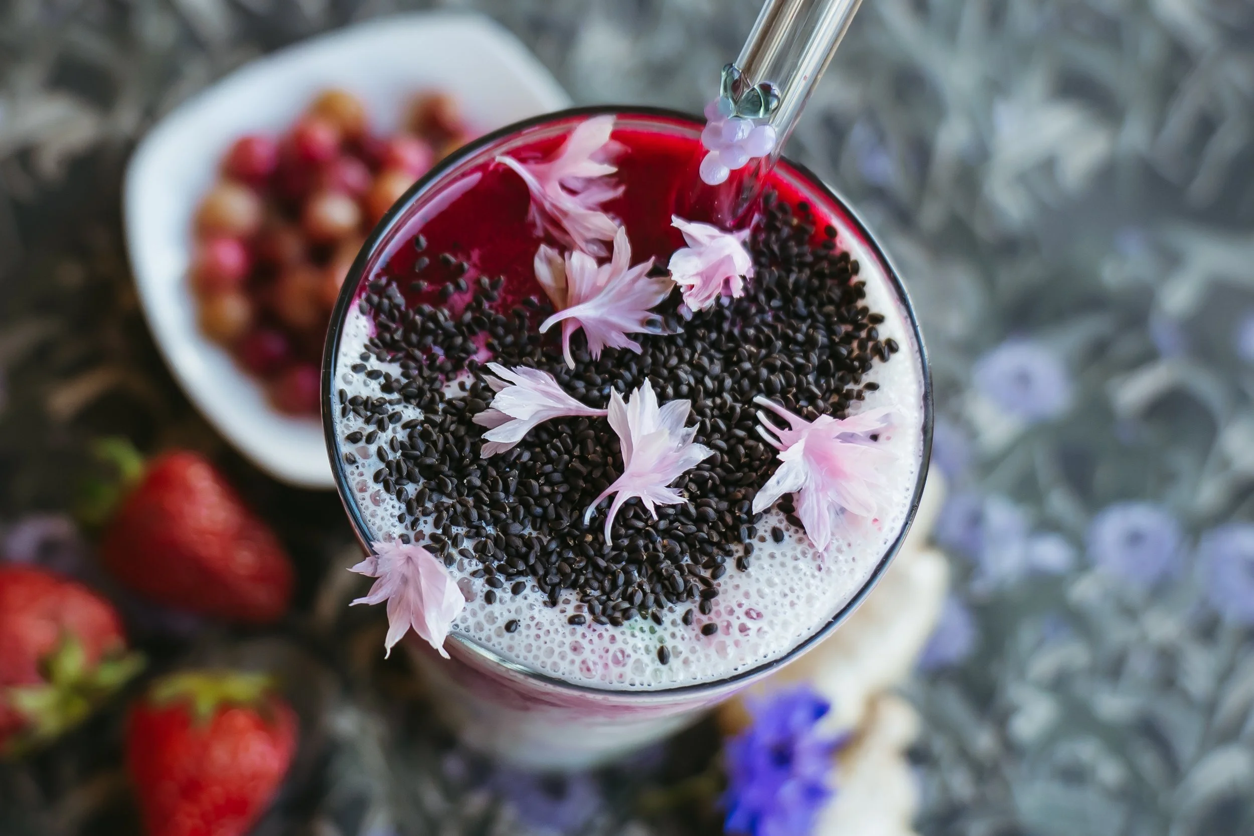Close-up of a pink beverage topped with black sesame seeds and pink flower petals, with a straw, surrounded by strawberries and other berries