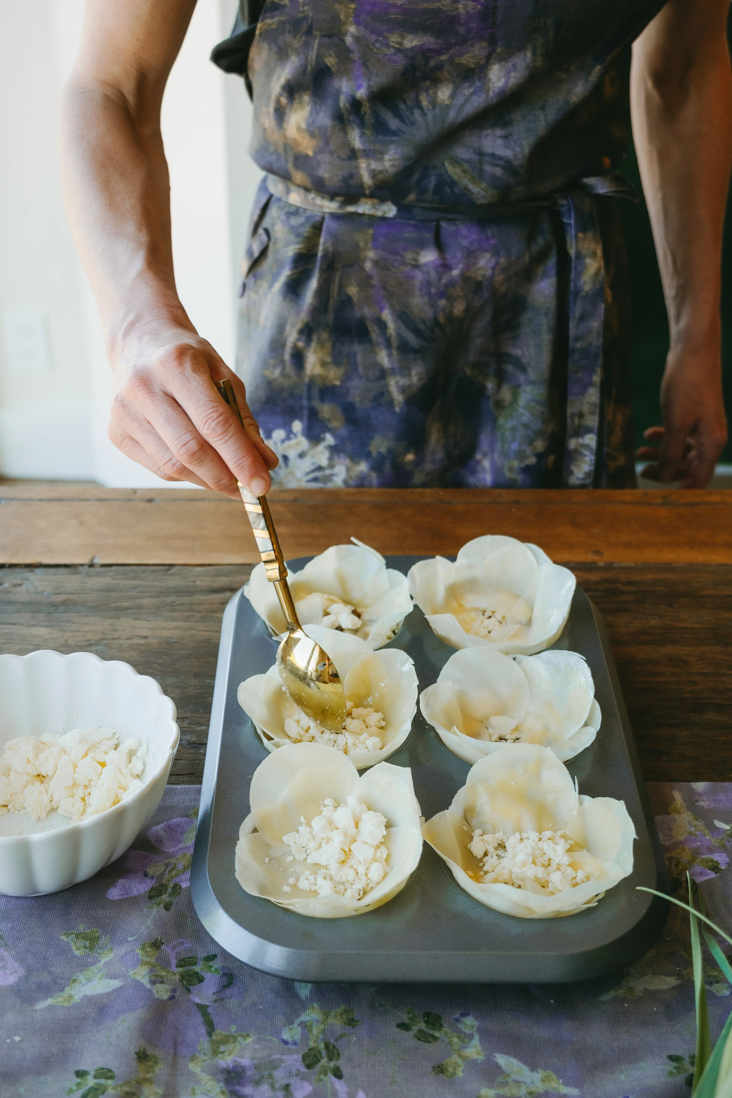 A person in a colorful dress is scooping filling into shell-shaped pastries on a tray, with bowls of filling on a table covered with a floral cloth.