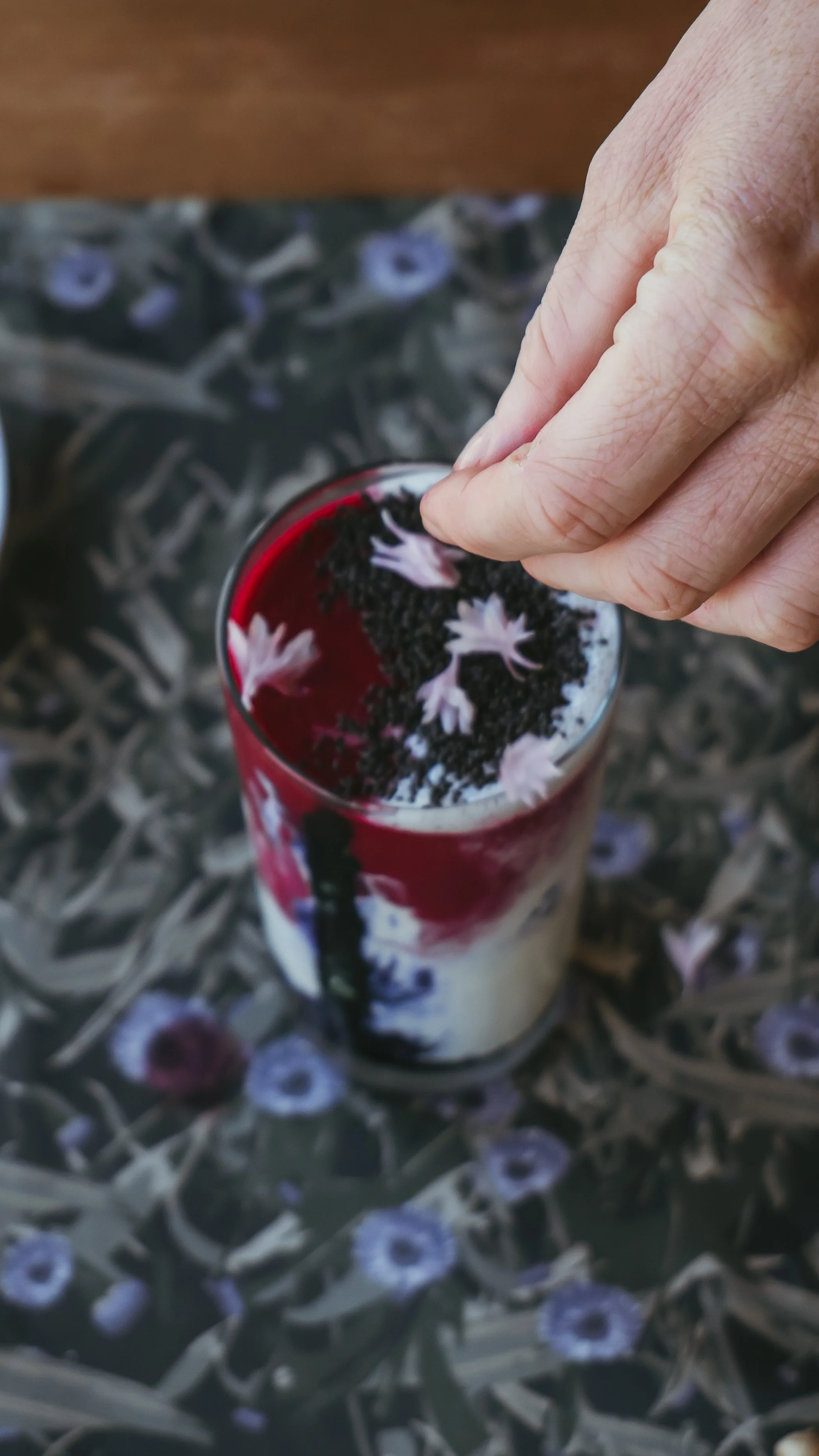 Close-up of a hand garnishing a layered berry dessert with edible flowers on top, seen from above.