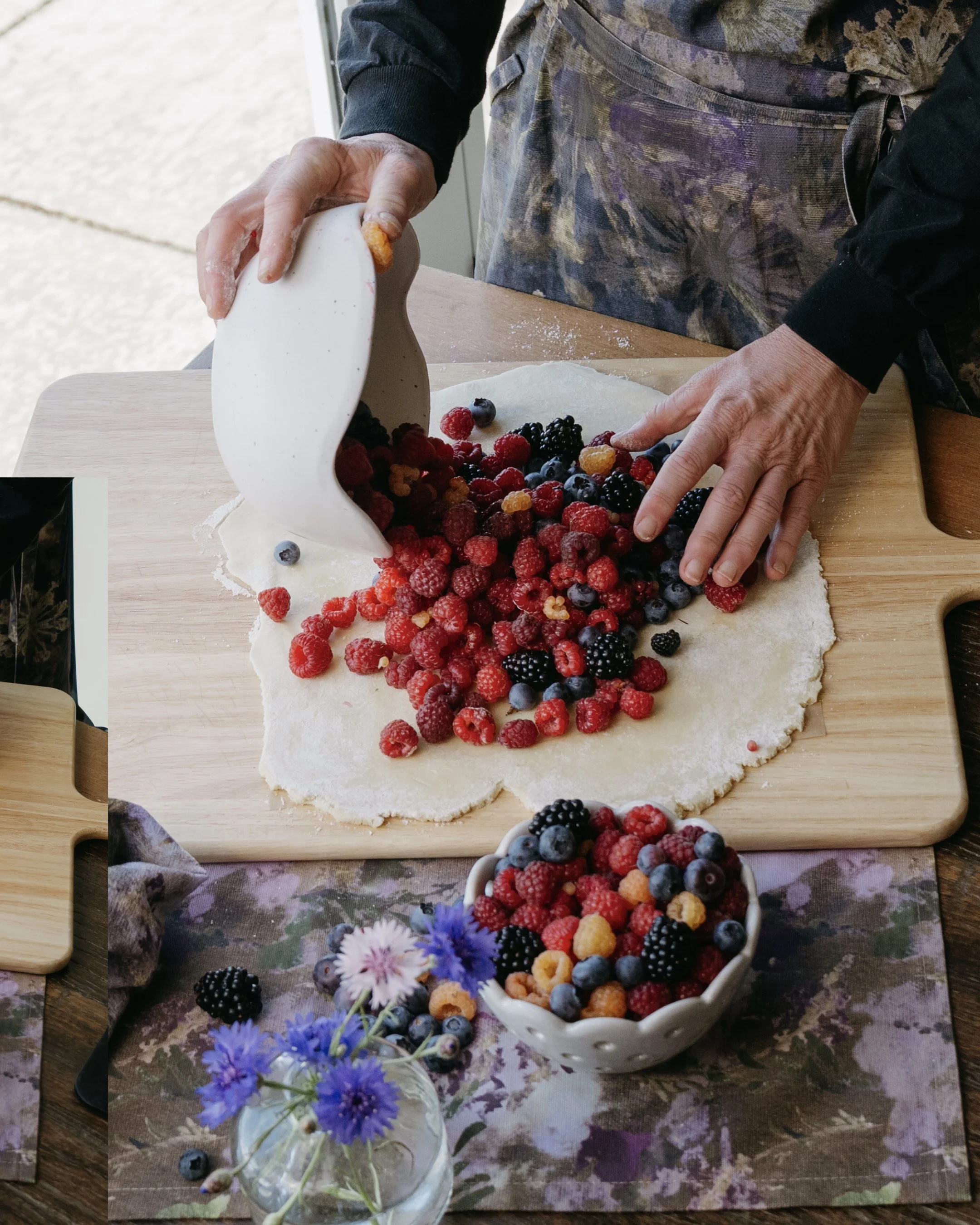 Snacky Girl is pouring fresh mixed berries onto a sheet of rolled-out dough, preparing to make a mixed berry galette. A bowl of berries and spring flowers decorates the table.