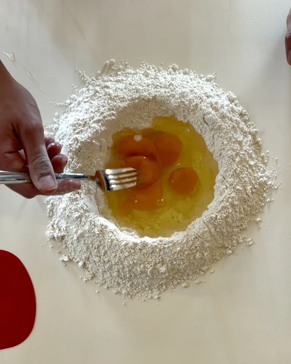 Bowl of flour with five raw eggs cracked into the center, with someone holding a fork ready to mix.