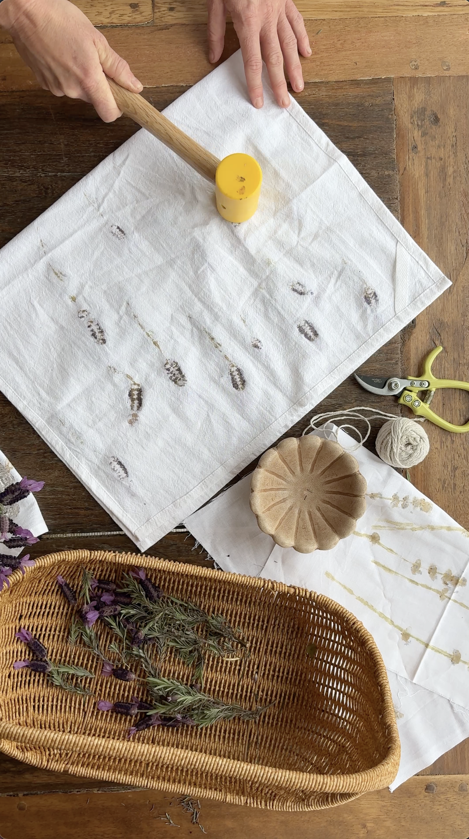 A person using a rubber mallet on a white cloth with dried lavender or similar herbs. The table has craft tools, including scissors, twine, a ceramic bowl, and a woven basket with sprigs of lavender or purple herbs.