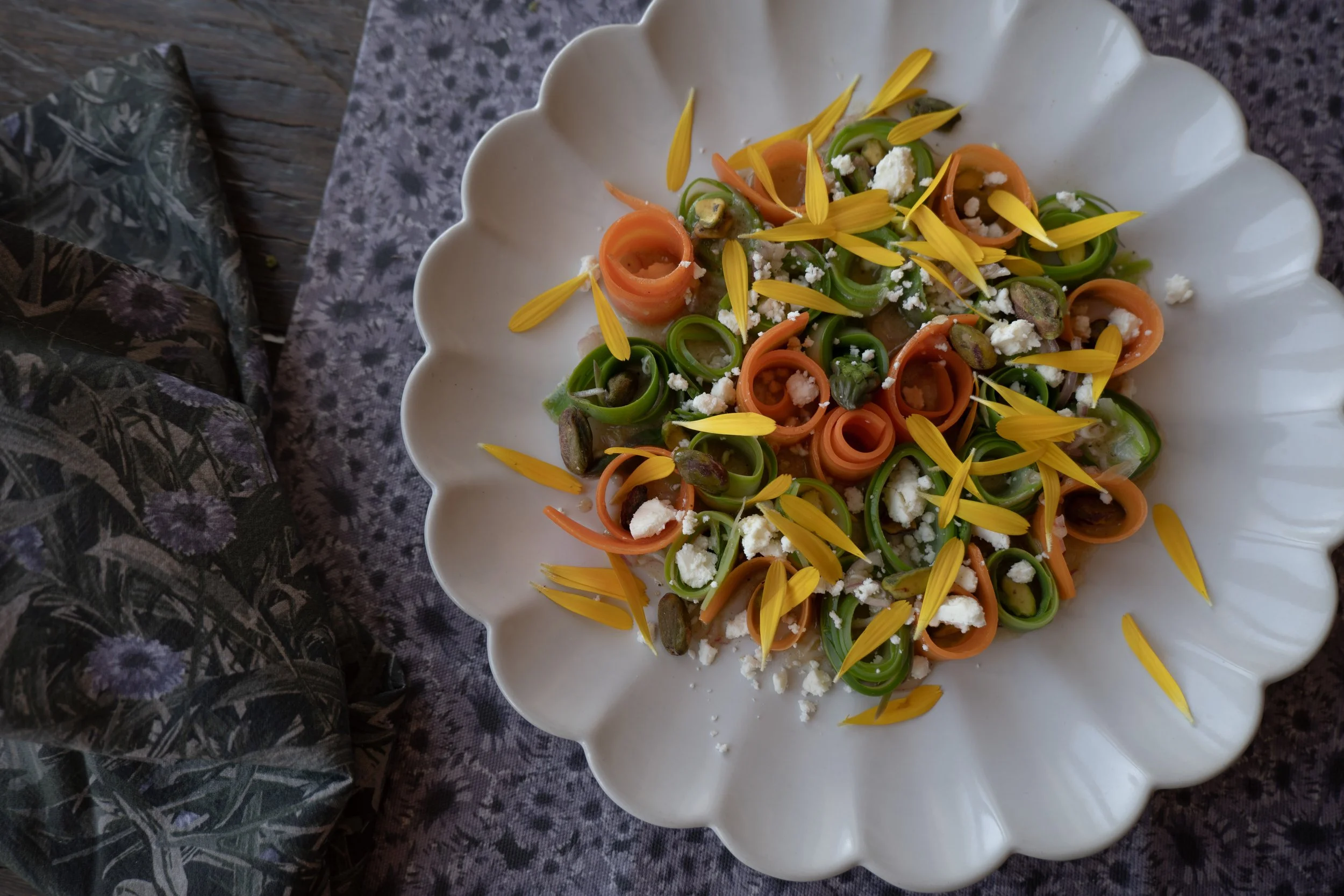 A white, scalloped-edge ceramic plate with colorful spiraled vegetable pasta, crumbled cheese, and yellow flower petals on a dark, speckled table.