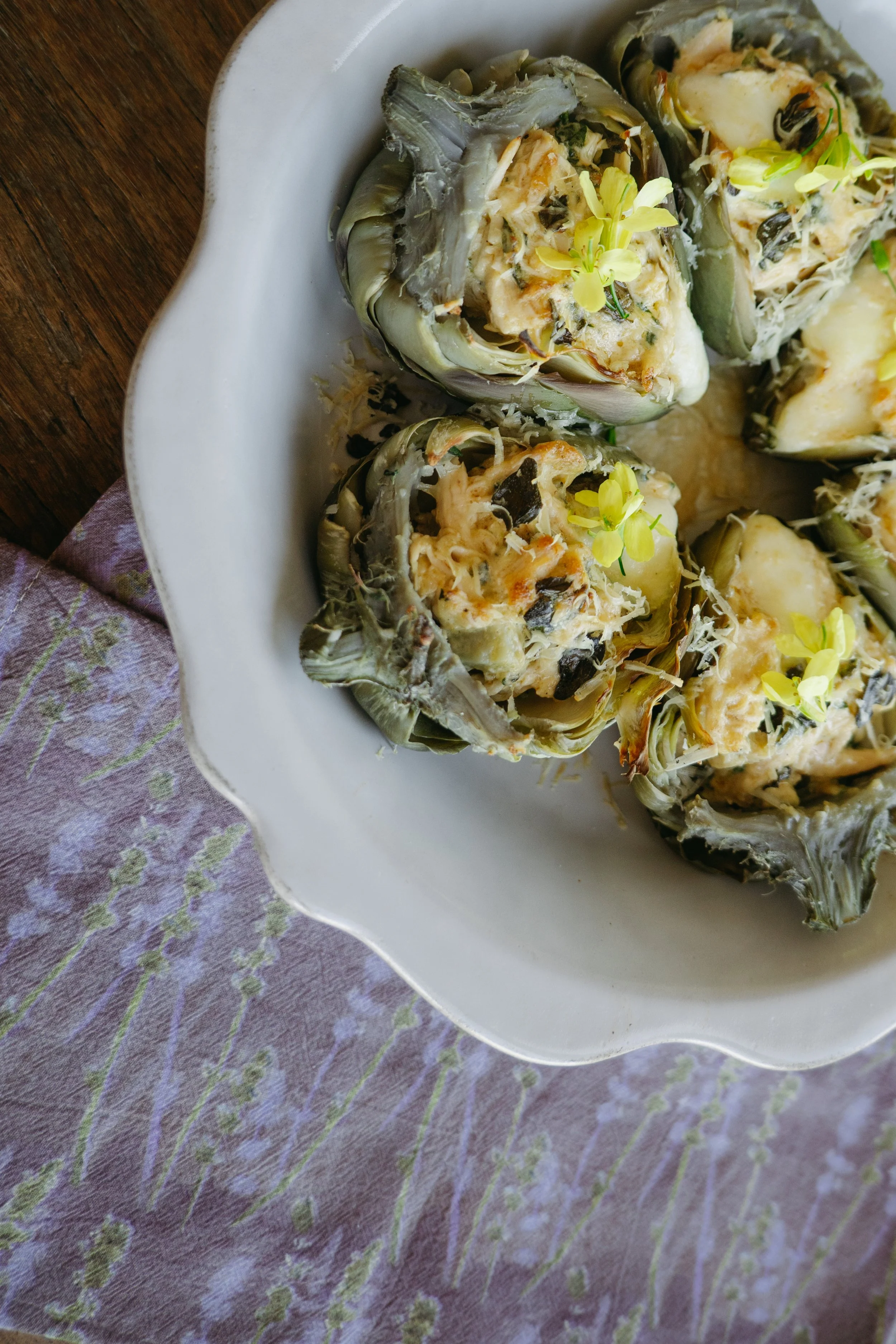 Decorative baked stuffed artichokes garnished with yellow edible flowers on a white serving dish placed on a purple fabric with green floral pattern.