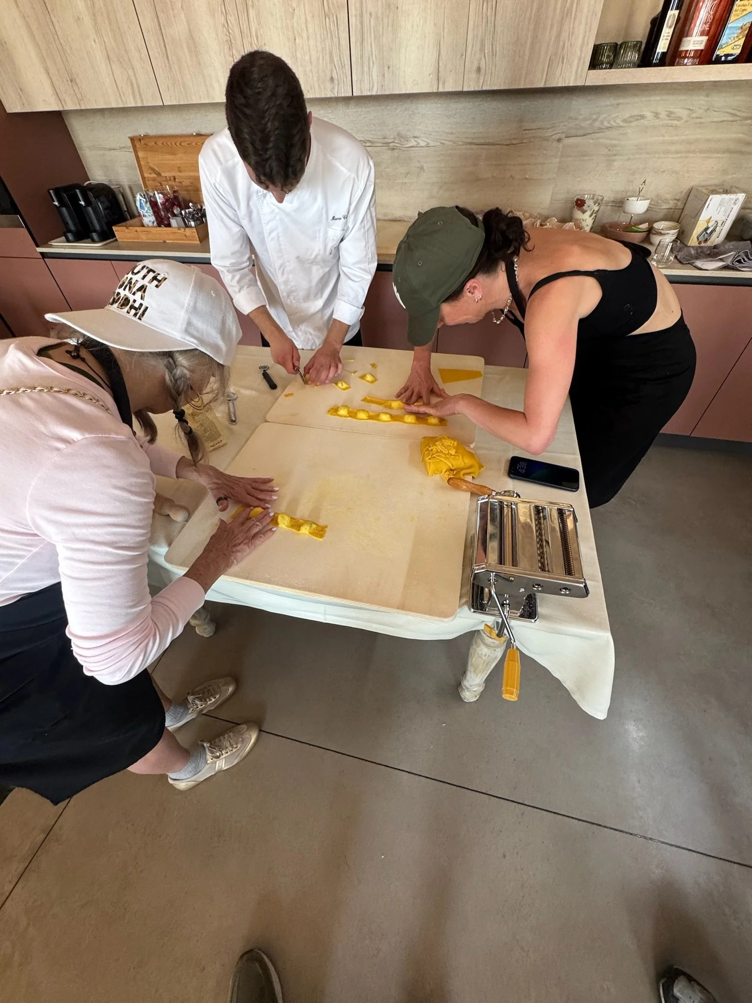 Three women are working together at a kitchen table, preparing yellow dough into strips. The women are focused on their tasks, with one wearing a large hat, another wearing a green cap, and the third in a white coat. The kitchen has pink cabinets and