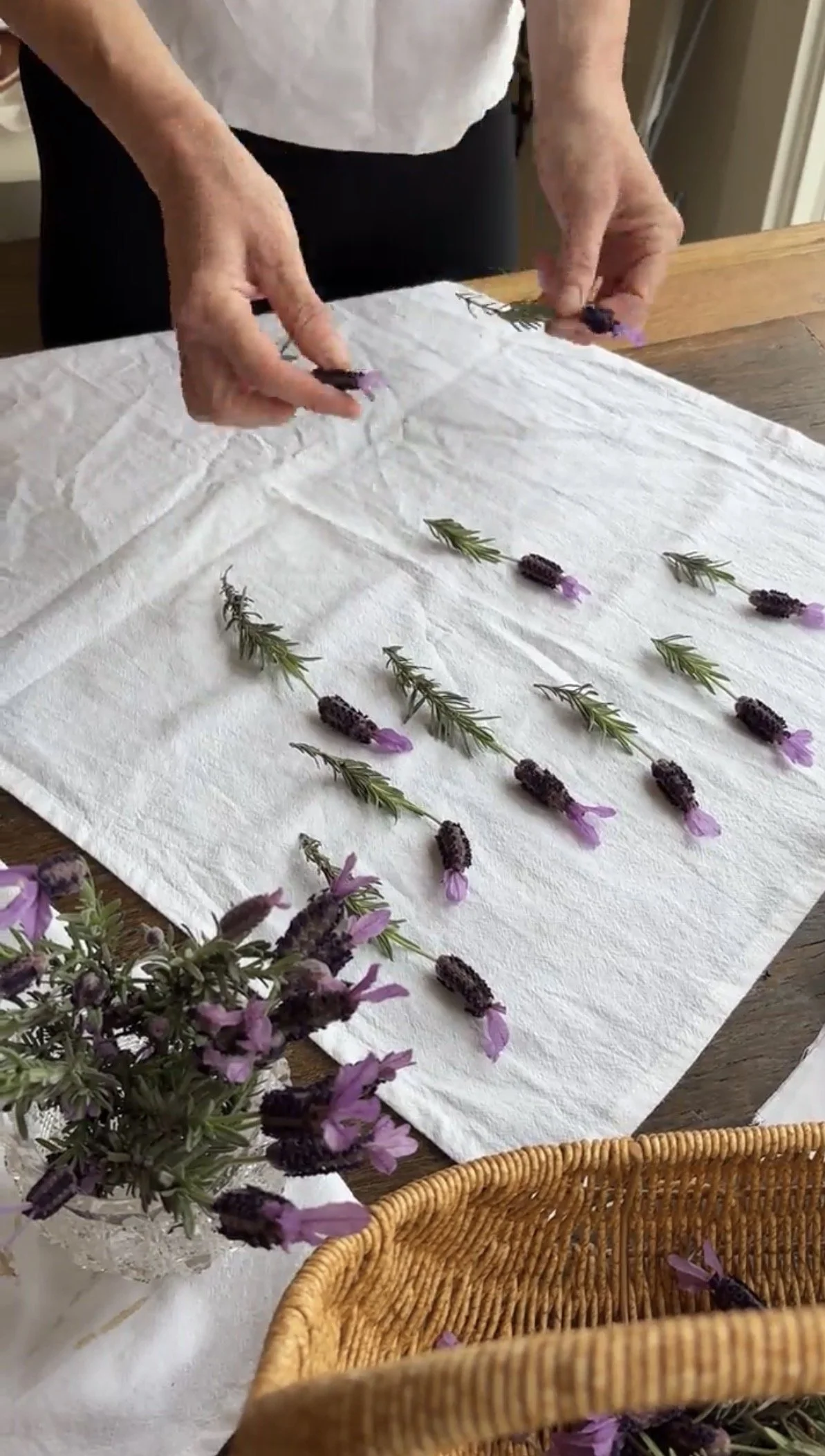 Hands arranging purple lavender flowers on a white cloth, with a basket and a purple flower bouquet nearby.