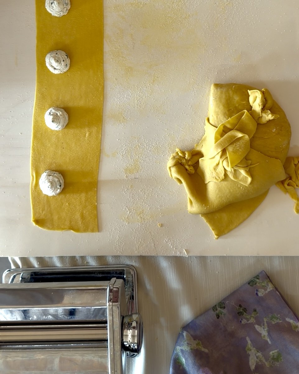 A sheet of yellow pasta dough with four dollops of white filling and a large piece of rolled-out dough on a kitchen counter.