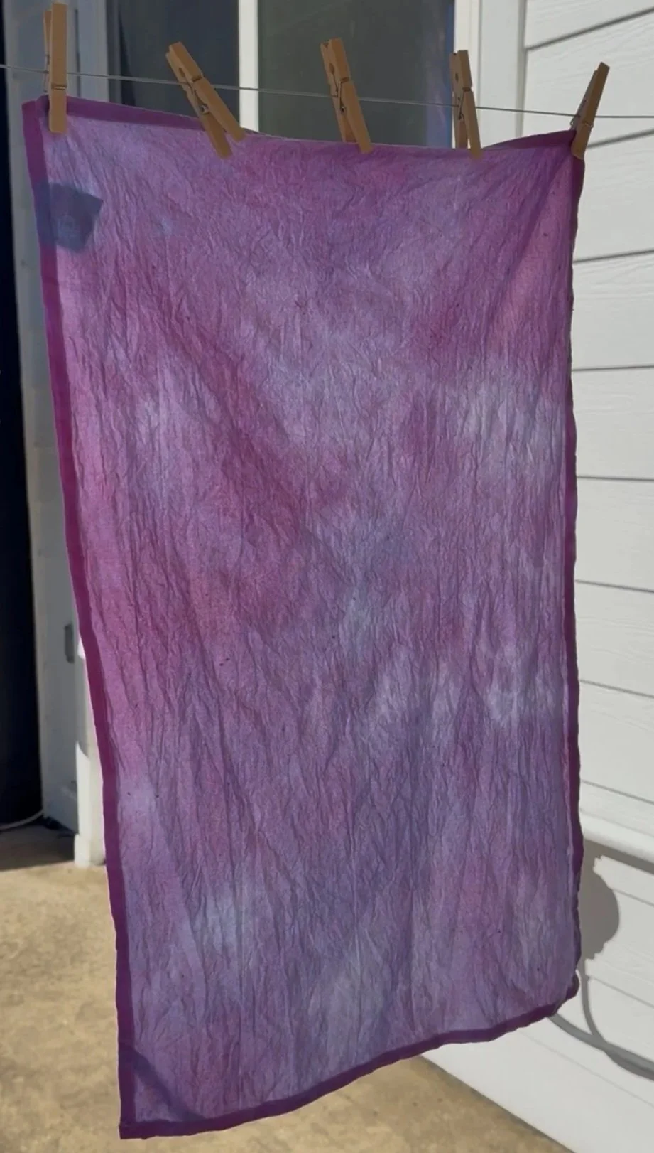A purple and pink fabric sheet hanging on a clothesline with three wooden clothespins, outside near a white building wall.