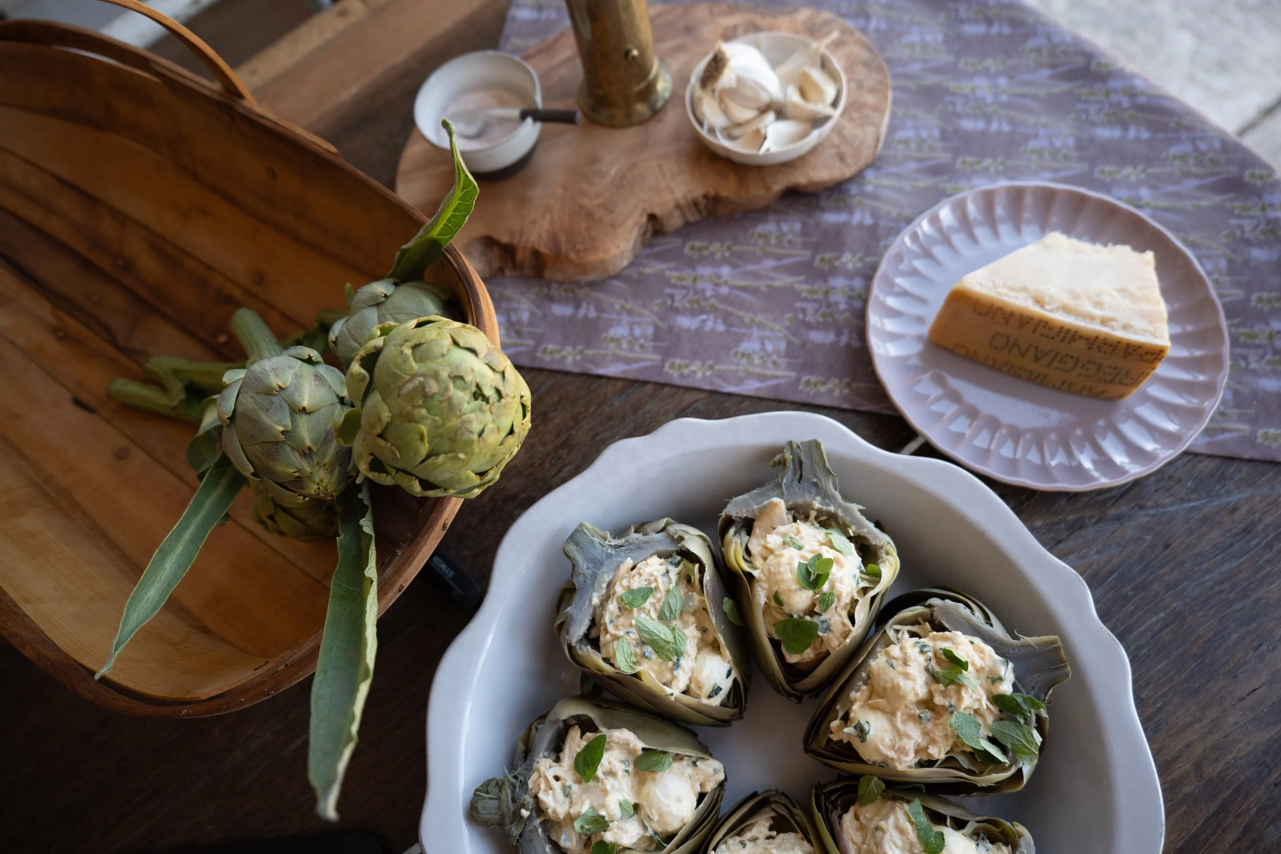 A wooden table with a white bowl of stuffed artichoke halves garnished with herbs, an artichoke, and a wooden board with bowls of garlic cloves and a liquid, possibly vinegar, along with a wedge of Parmesan cheese on a pink paper plate, all arranged on a purple cloth.