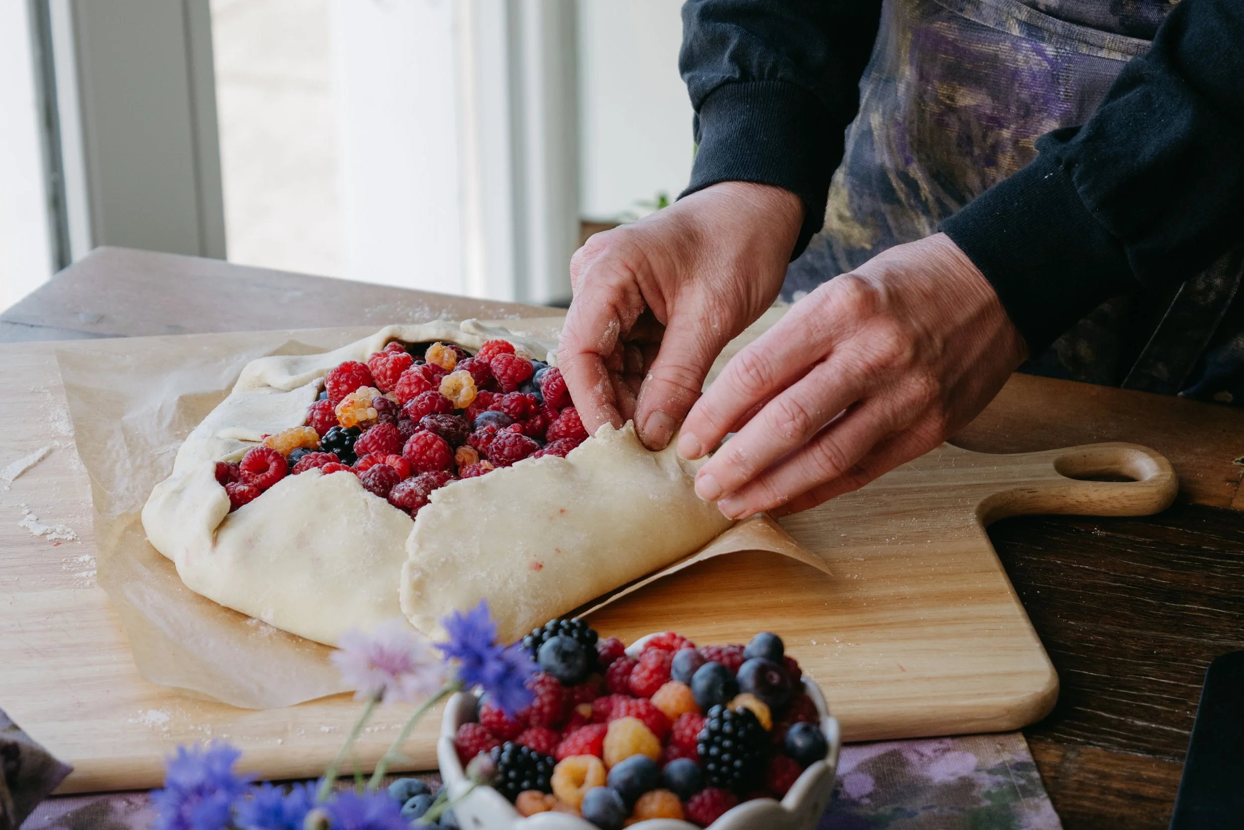Snacky Girl making a berry galette on a wooden table with fresh berries and flowers nearby.