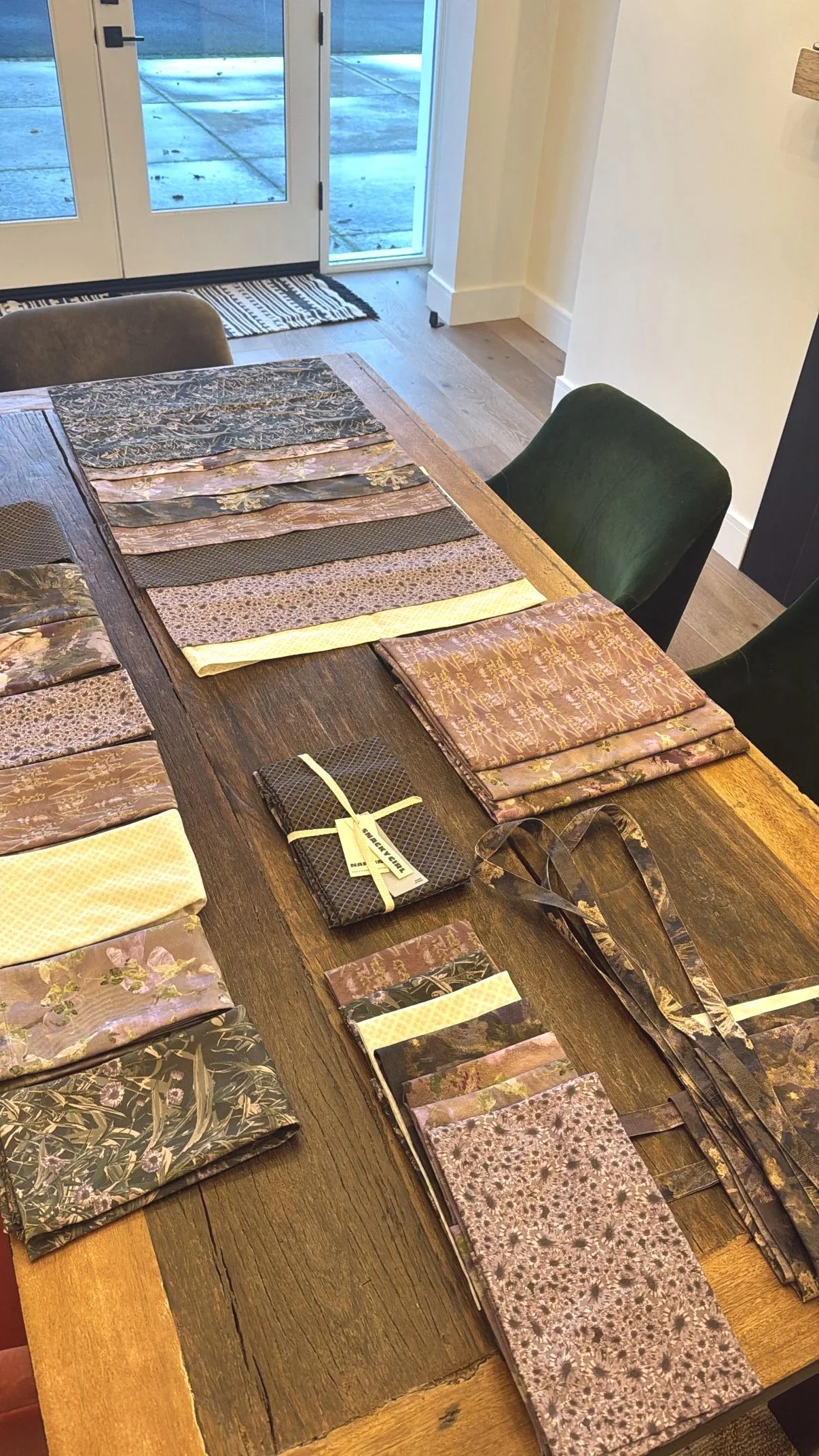 A wooden table displaying an array of folded fabrics with floral and patterned designs, and a pair of glasses, in a room with glass doors leading outside.