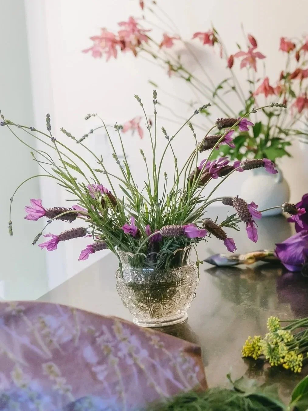 A clear glass vase filled with a mixed bouquet of purple and lavender flowers on a dark table. In the background, there are pink flowers in a white vase and some other colorful flowers on the table.