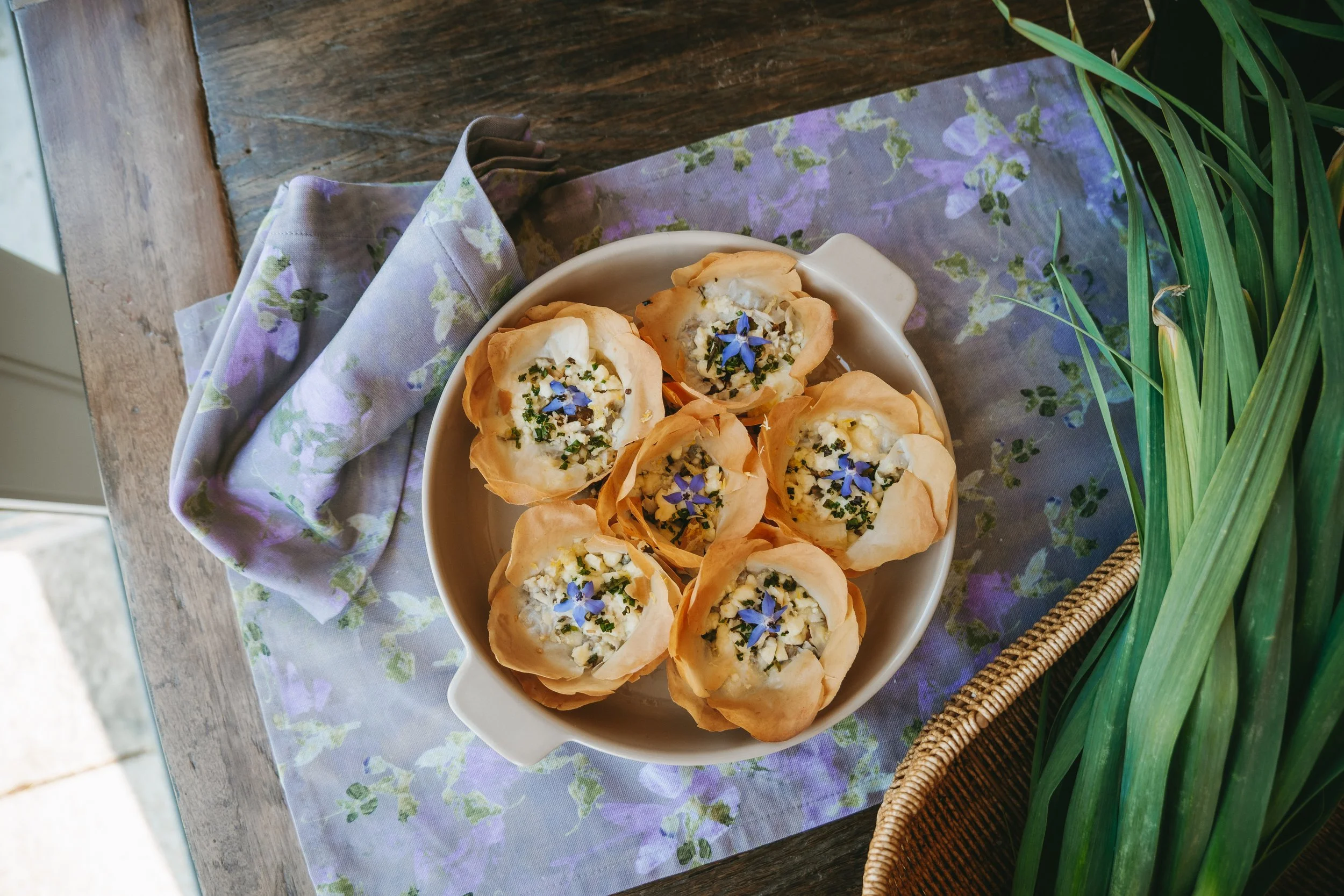 A white bowl filled with phyllo pastry cups topped with cheese, herbs, and edible flowers, placed on a purple floral napkin on a wooden table, with a basket of greenery nearby.