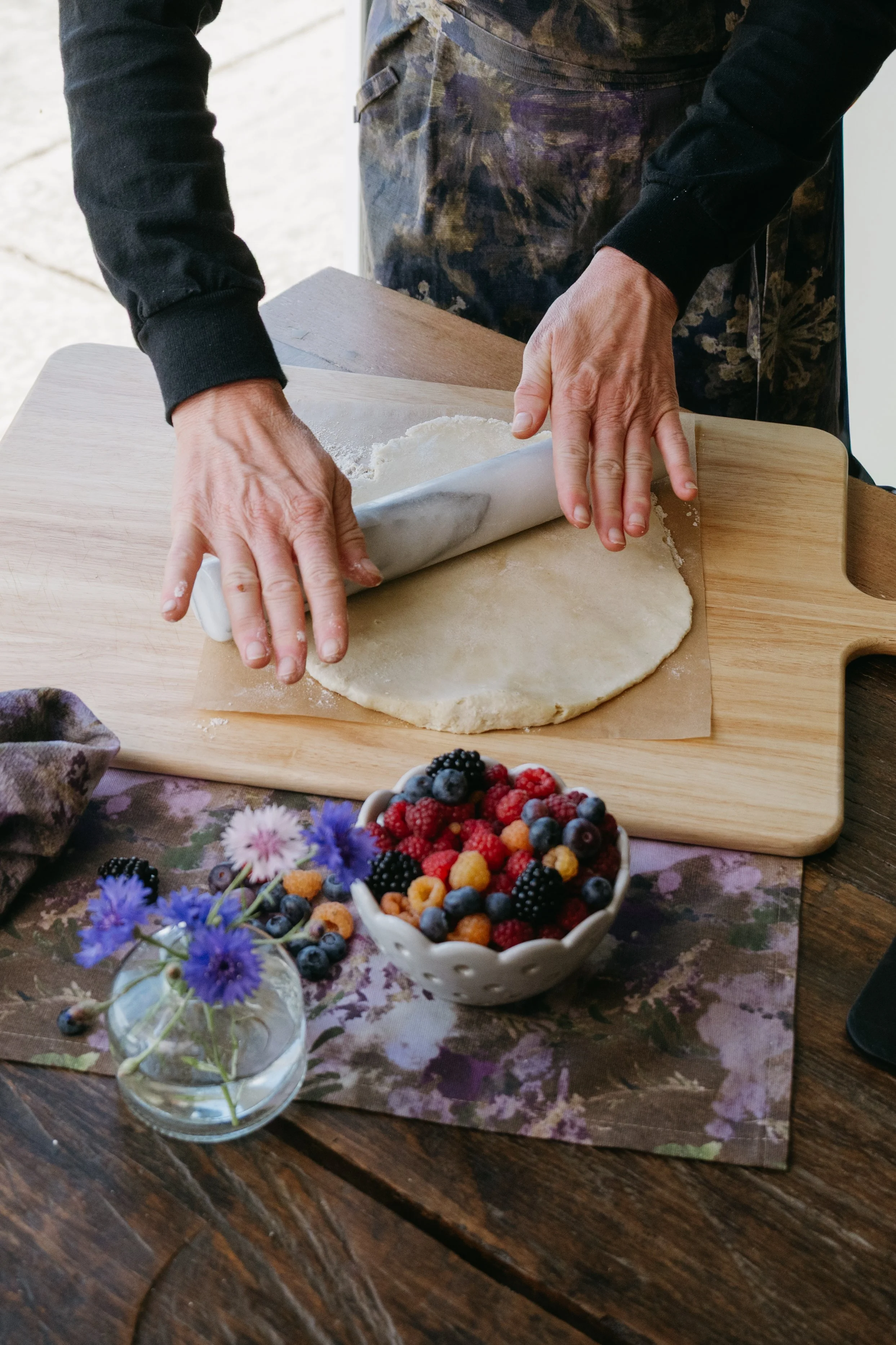 Snacky Girl rolling out dough on a wooden surface, with a bowl of mixed berries and a small vase of purple and blue flowers on the table.