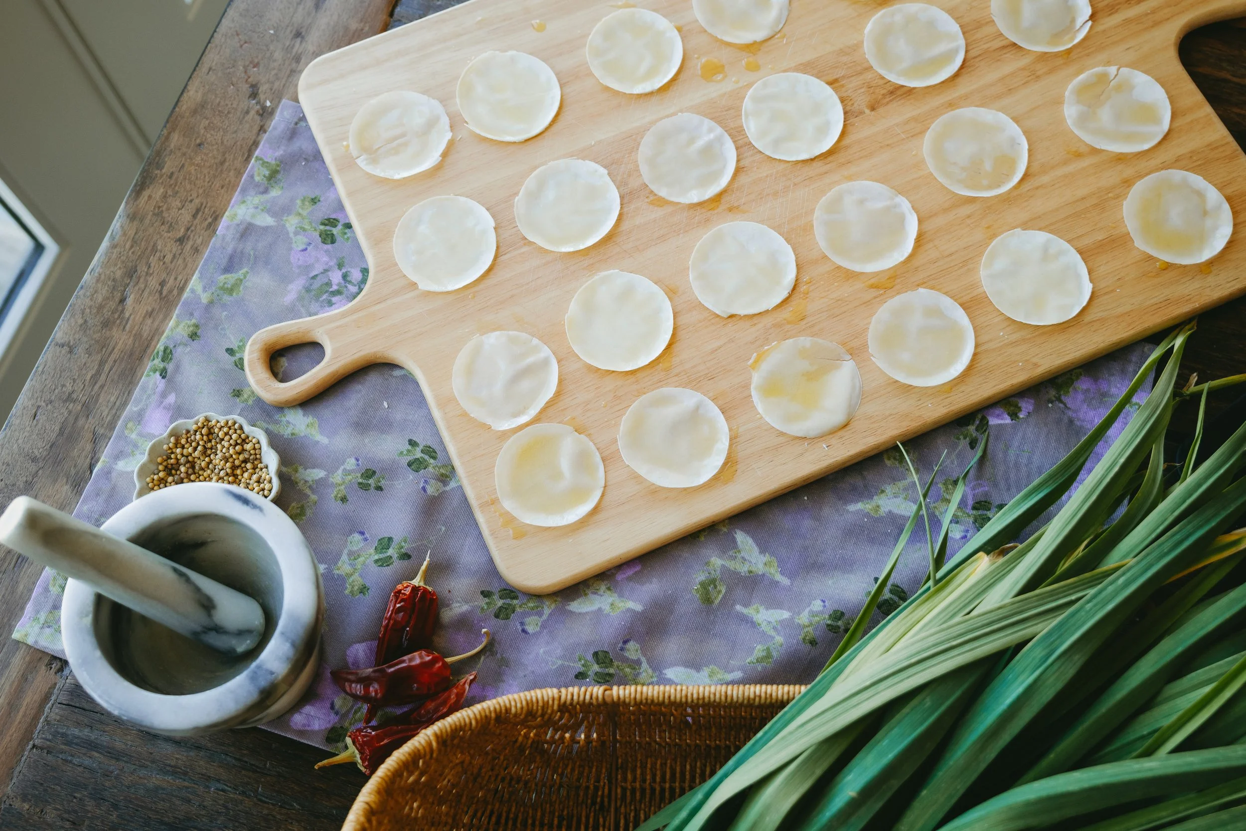 A wooden cutting board with circular dumpling wrappers arranged in rows. Nearby, a small bowl of whole peppercorns, some dried red chili peppers, and green onion stalks are visible, along with a purple floral cloth, a white mortar and pestle, and a w