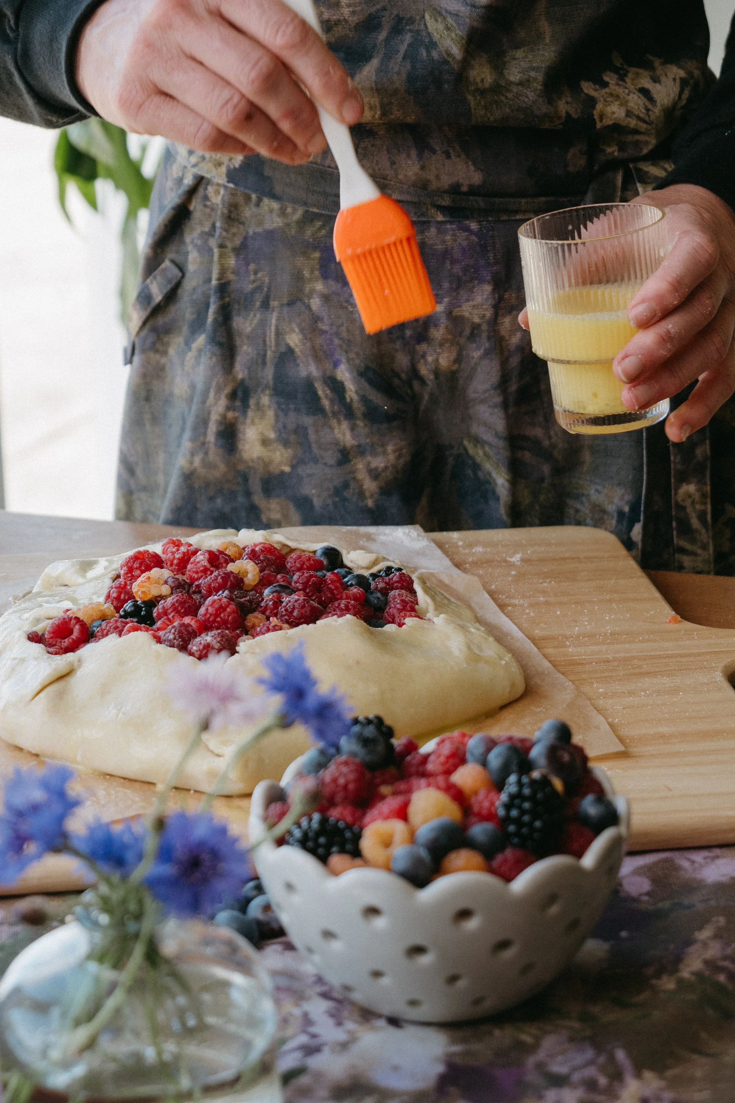 Snacky Girl pouring orange sauce or syrup on a fruit tart topped with various berries, with a bowl of mixed berries nearby and a vase with blue flowers in the foreground.