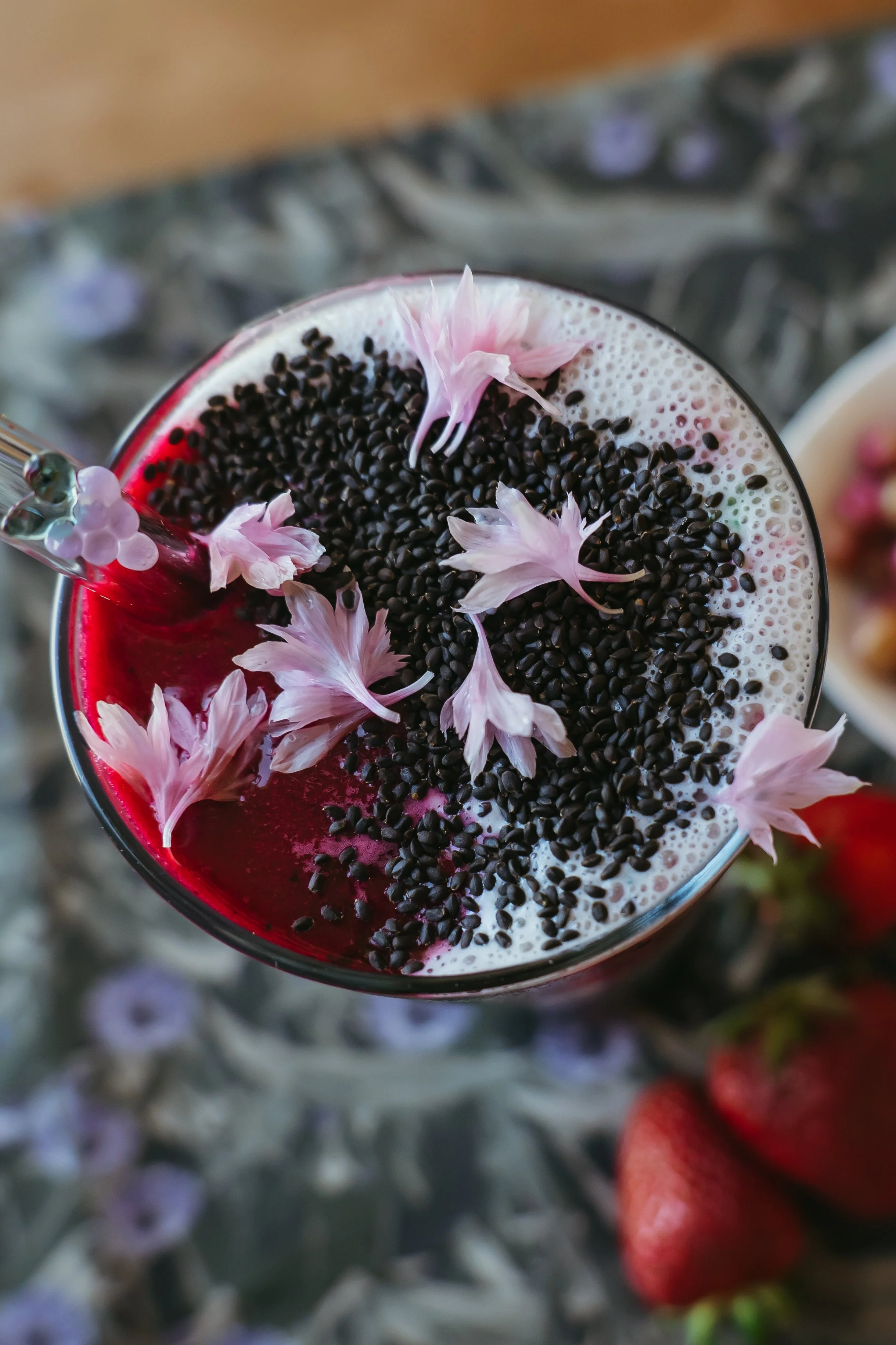 A glass of a pinkish red beverage topped with black sesame seeds and light pink flower petals, with a decorative straw, against a background of blurred flowers and strawberries.