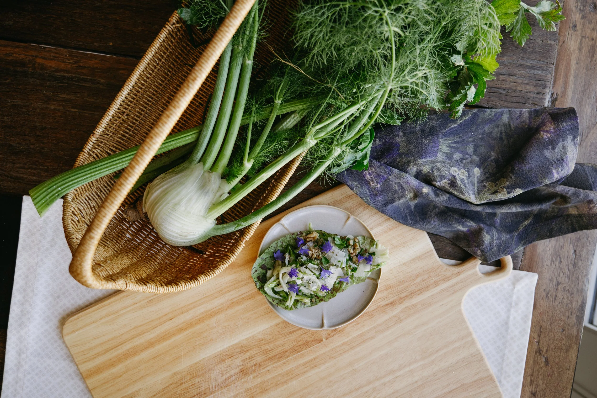 A basket with fresh green vegetables, including spring onions and leafy greens, placed on a wooden table, with a small bowl of salad nearby.