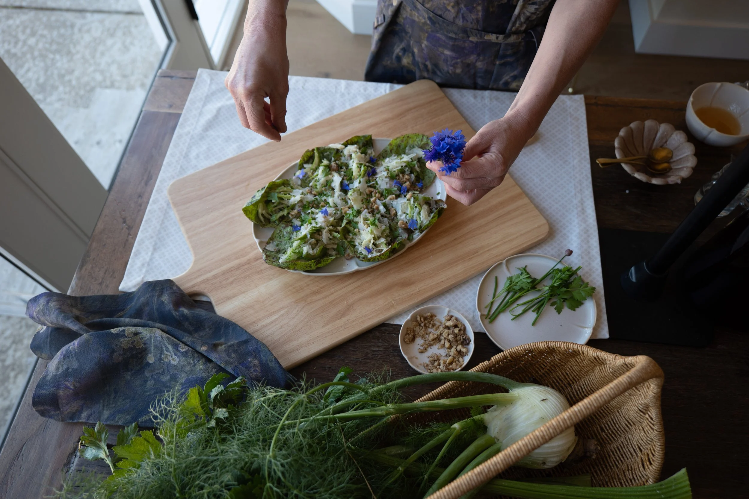 A person preparing a fresh salad with lettuce, cheese, and edible blue flowers on a white plate, surrounded by various ingredients and herbs on a wooden table in a bright kitchen.