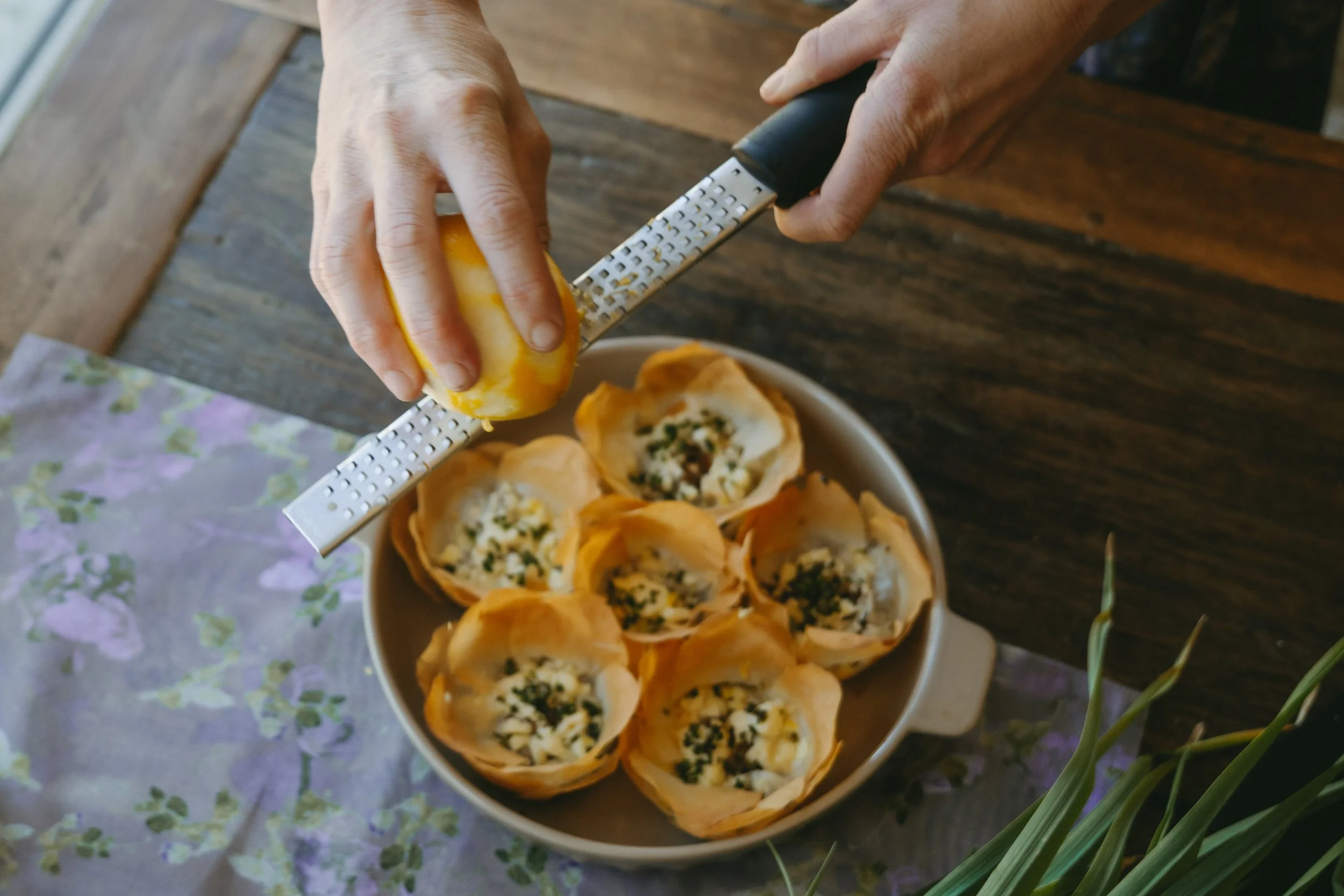 Person grating a lemon over a bowl of baked pastry cups filled with cheese and herbs on a wooden table.