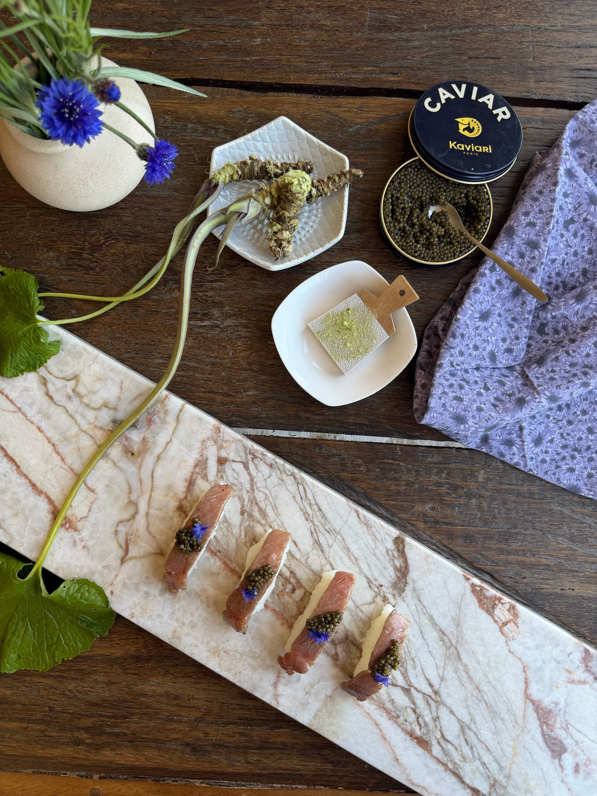 Sushi rolls topped with caviar, garnished with small purple flowers, arranged on a marble slab. A small bowl of caviar with a spoon, a white dish with green powder, a sprig of greenery, and a Victorian-style cloth are also visible on a wooden table.