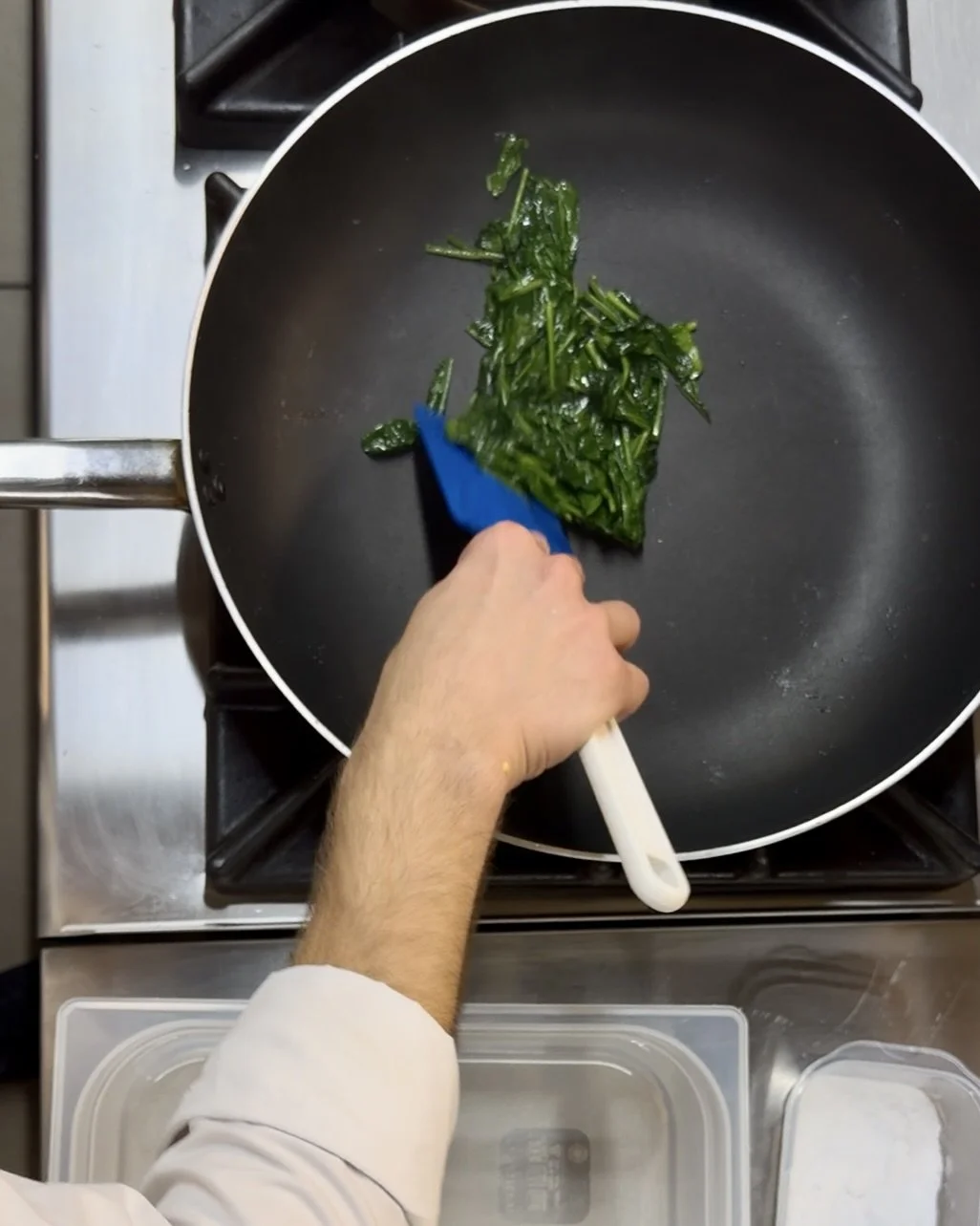 Person cooking spinach in a black skillet on a stove, using a spatula.