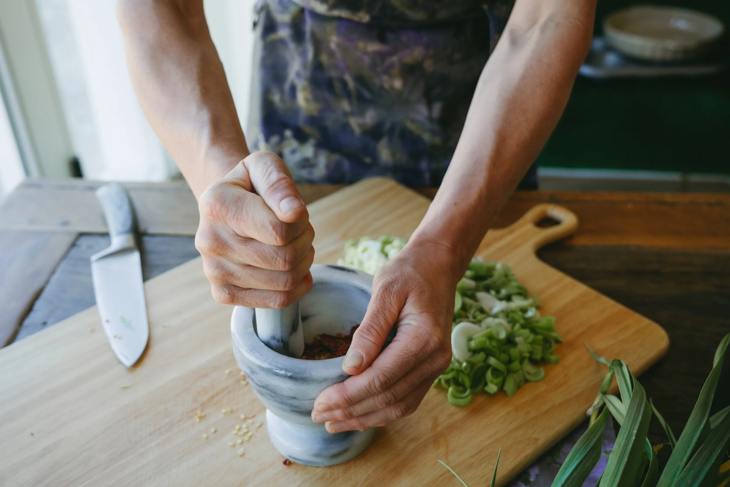 Person using a mortar and pestle to grind spices on a wooden cutting board with chopped vegetables and herbs nearby.