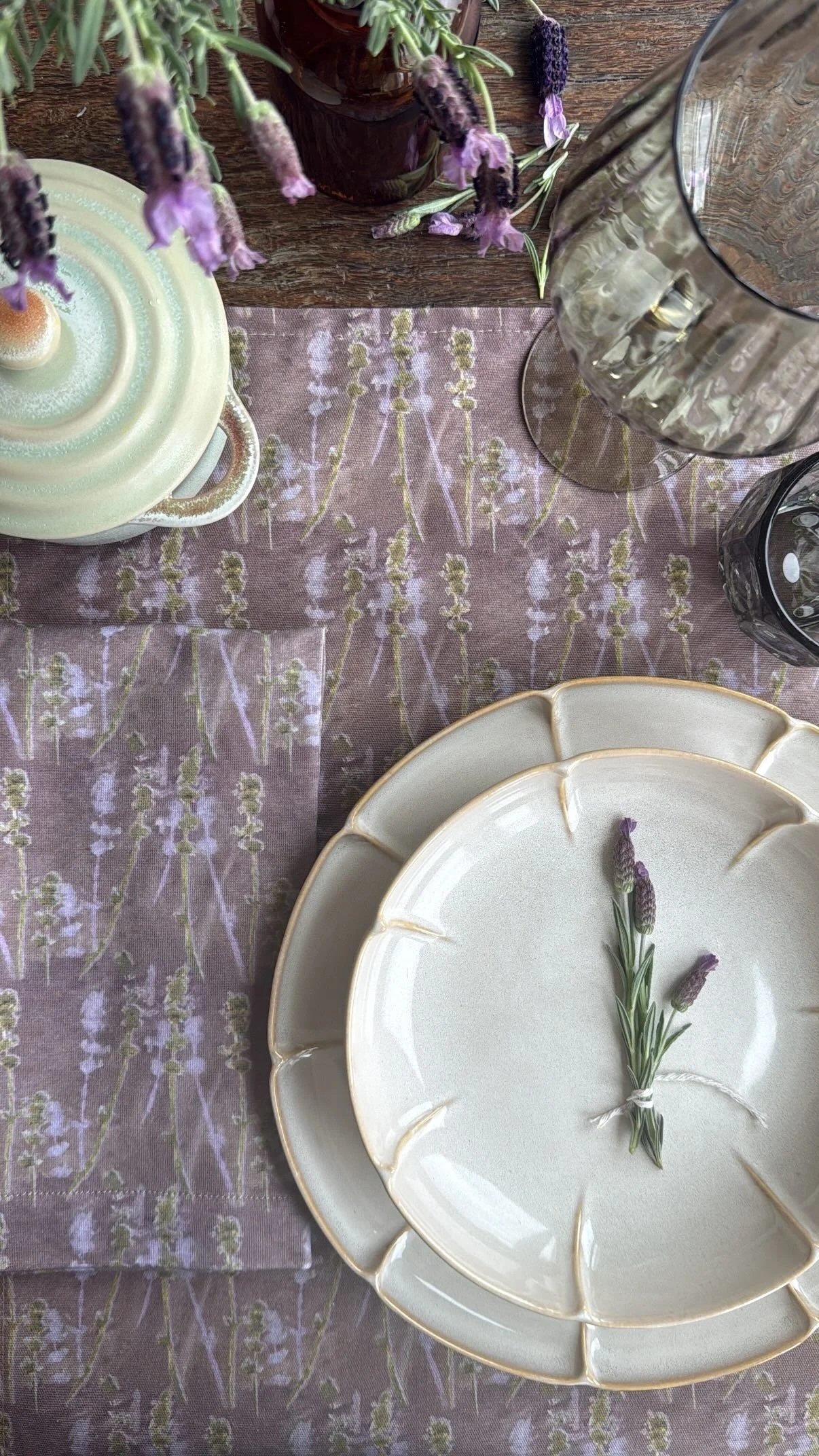 A table setting with a lavender tablecloth, white ceramic plates with gold edges, a sprig of lavender tied with string, glasses, a candle, and lavender flowers, with a wooden surface and potted lavender in the background.