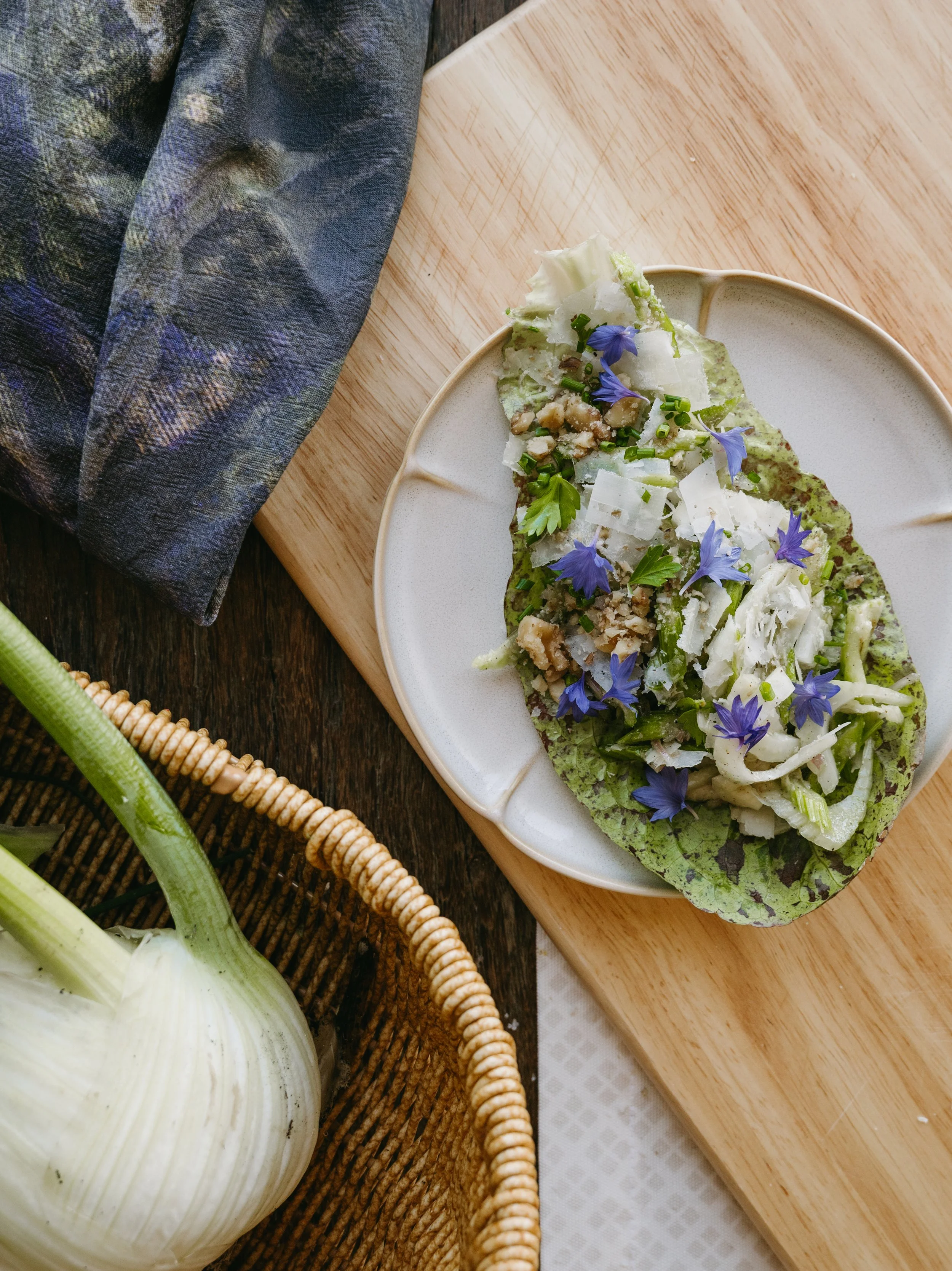 A dish of stuffed endive leaves garnished with edible flowers, chopped nuts, and shaved cheese on a white plate, placed on a wooden cutting board.