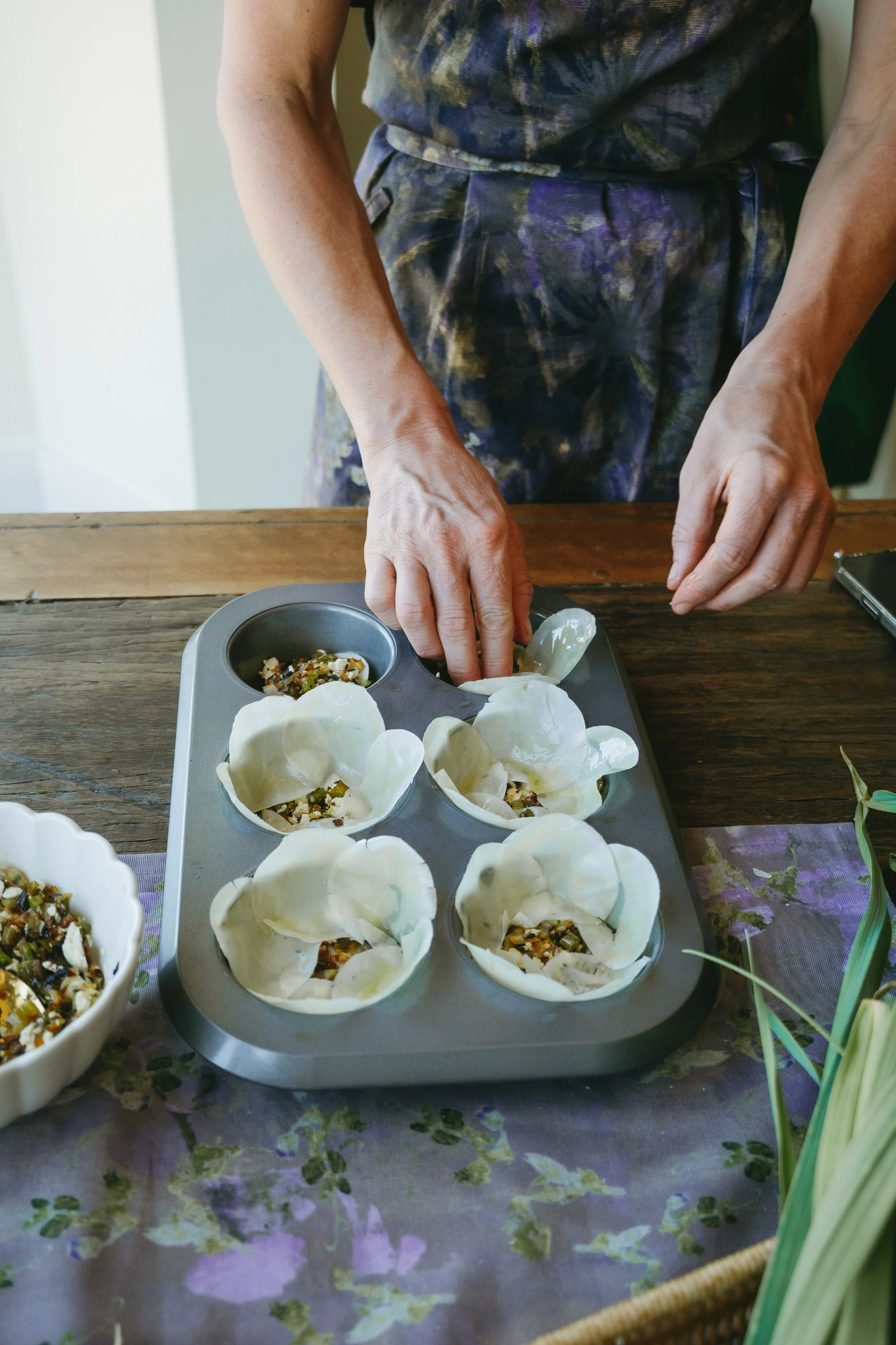 Person filling small pastry shells with a savory filling from a bowl, with a baking tray of prepared shells on a table.