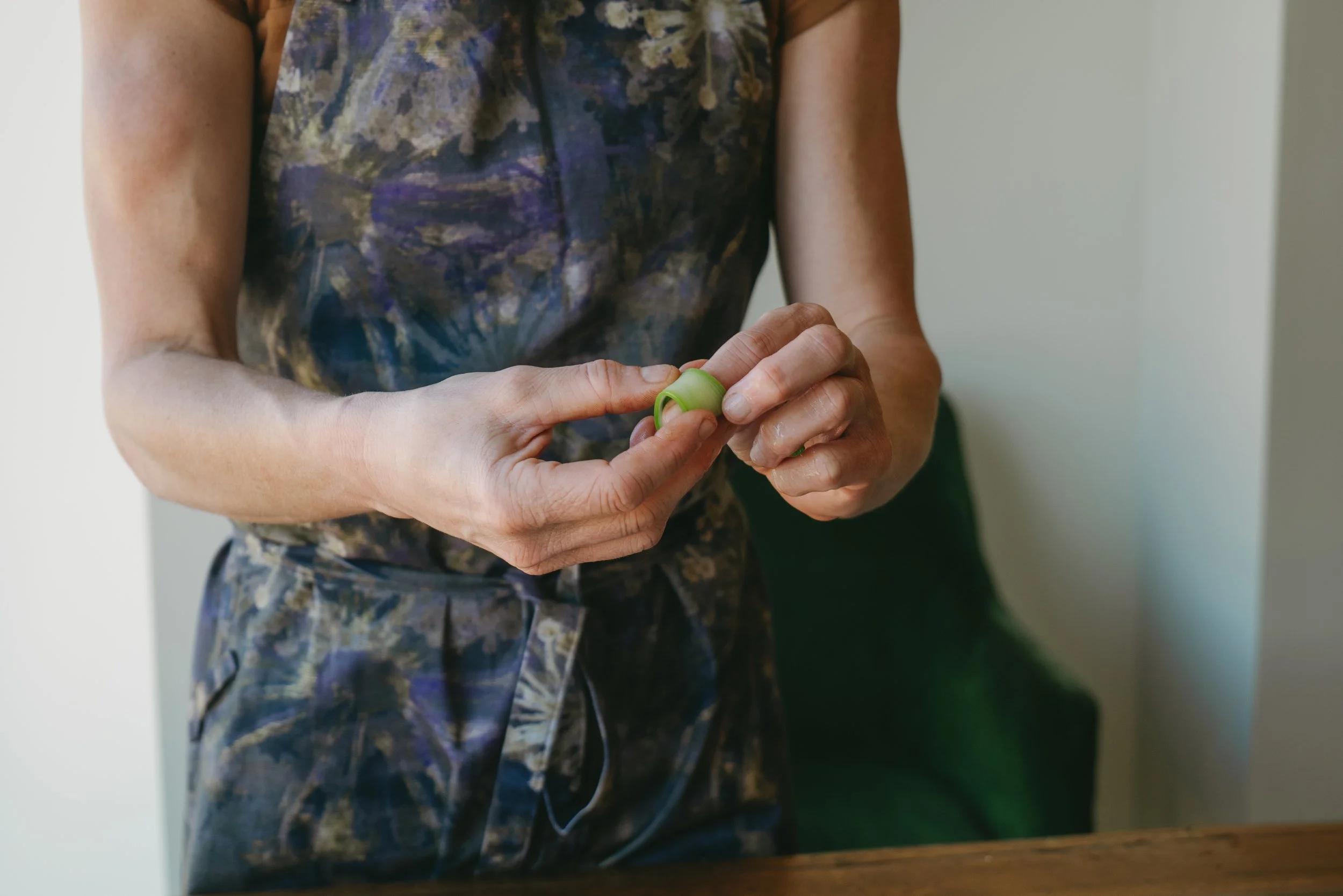 Close-up of a woman holding a small green object or tool in her hands. She is wearing a colorful, patterned sleeveless top and matching pants. The background is a simple indoor setting with off-white walls.