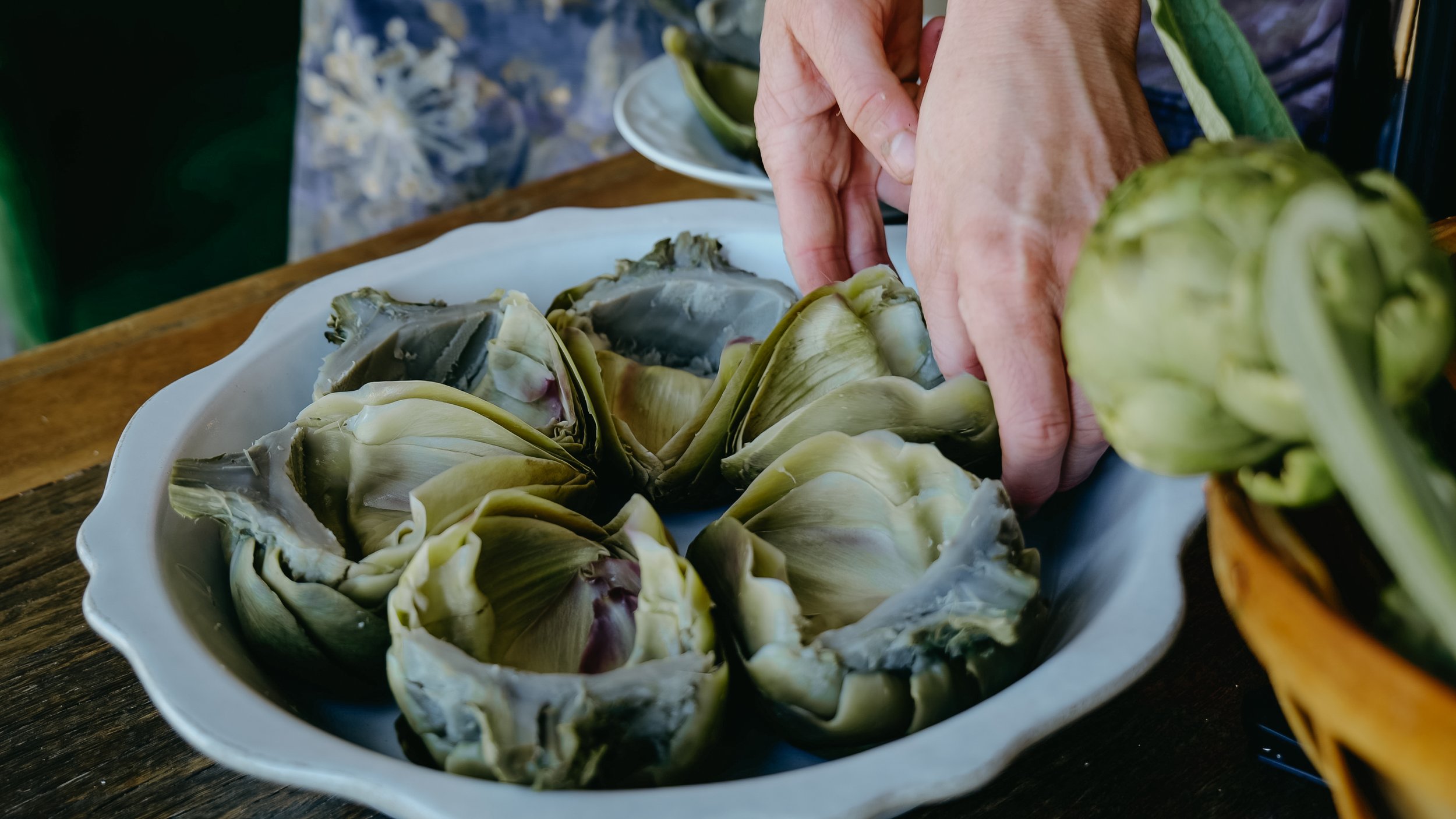 Hands preparing sliced artichokes on a white plate, with a lime wedge and other dishes in the background.
