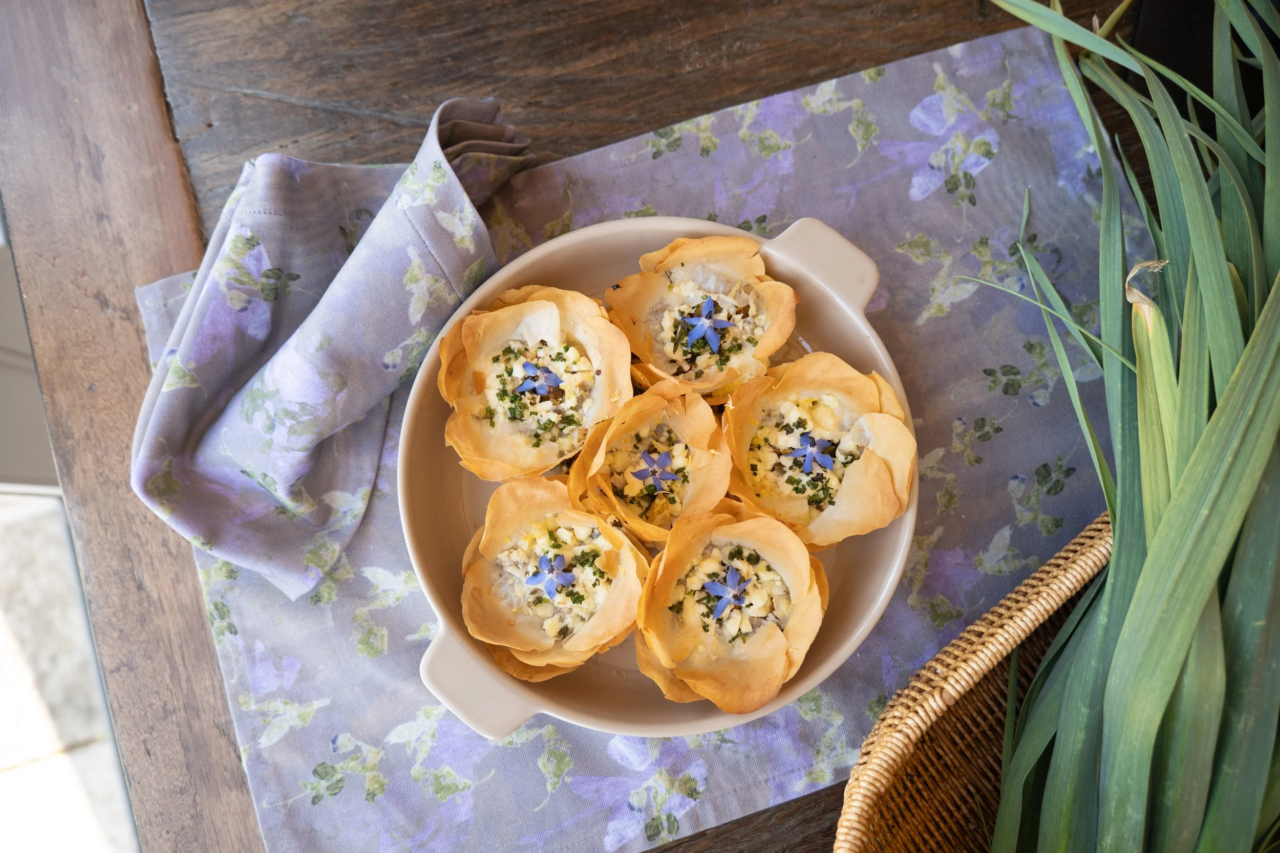 A white bowl filled with small savory pies topped with chopped herbs and blue edible flowers, placed on a table with a floral cloth and surrounded by green plants.