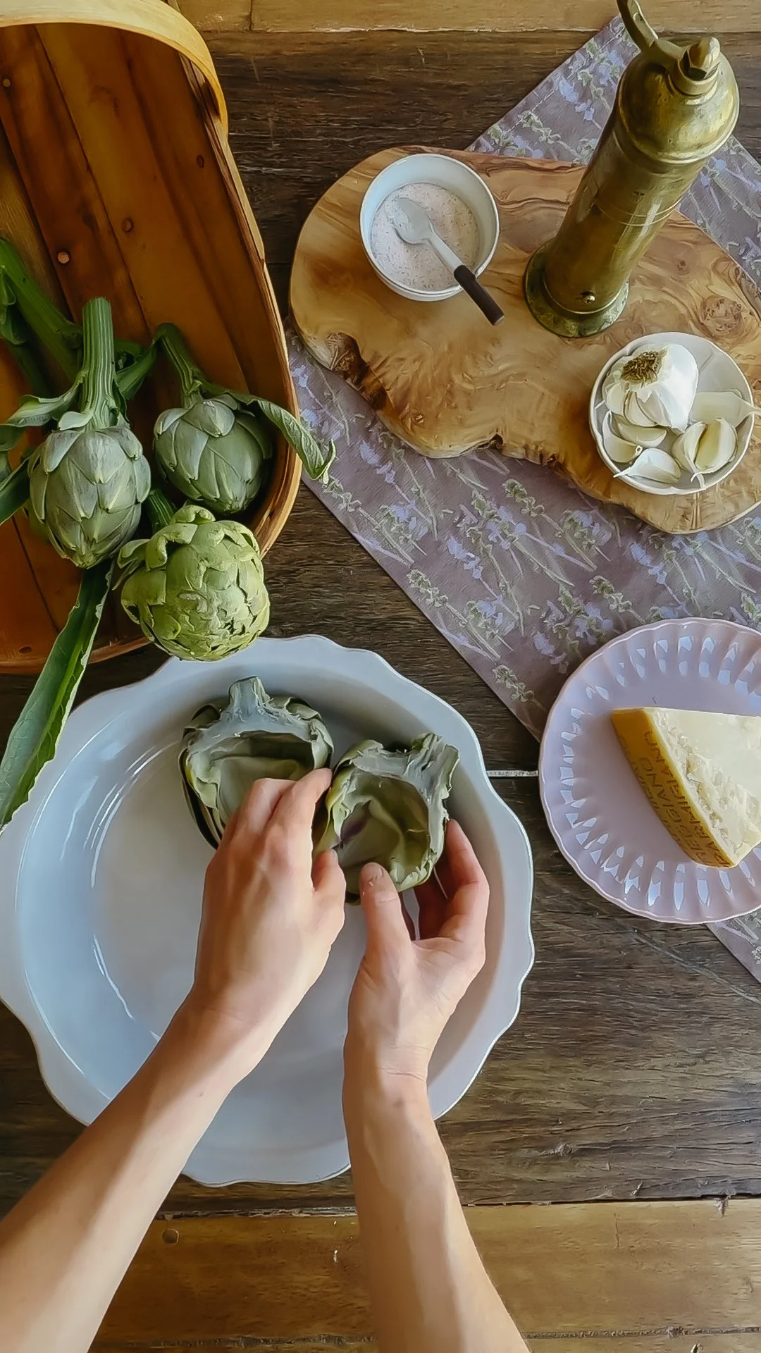 A person preparing fresh artichokes on a white plate at a wooden table, with fresh artichokes in a basket, a bowl of cheese, and a jar of oil on a wooden cutting board.