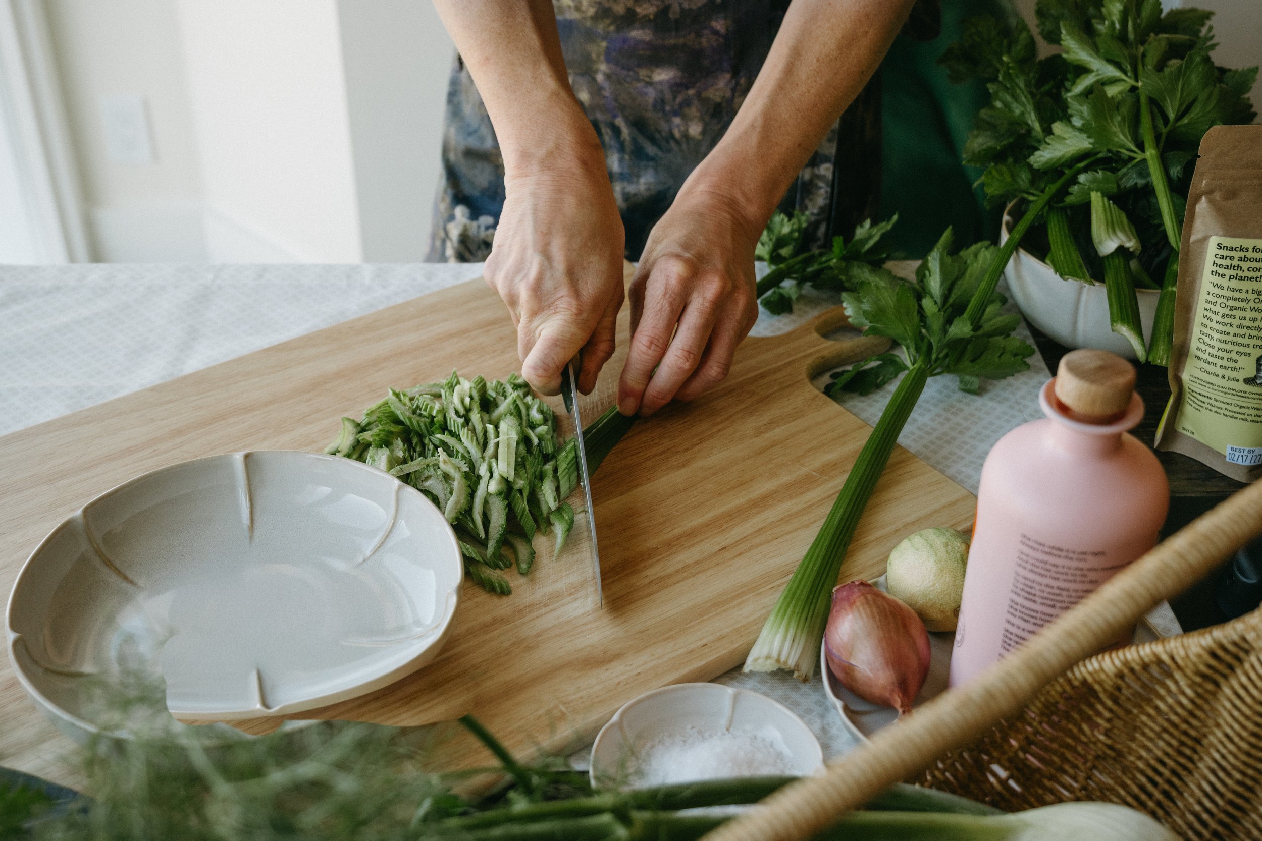 Person chopping celery on a wooden cutting board in a kitchen, surrounded by fresh vegetables, a bowl, and other kitchen items.