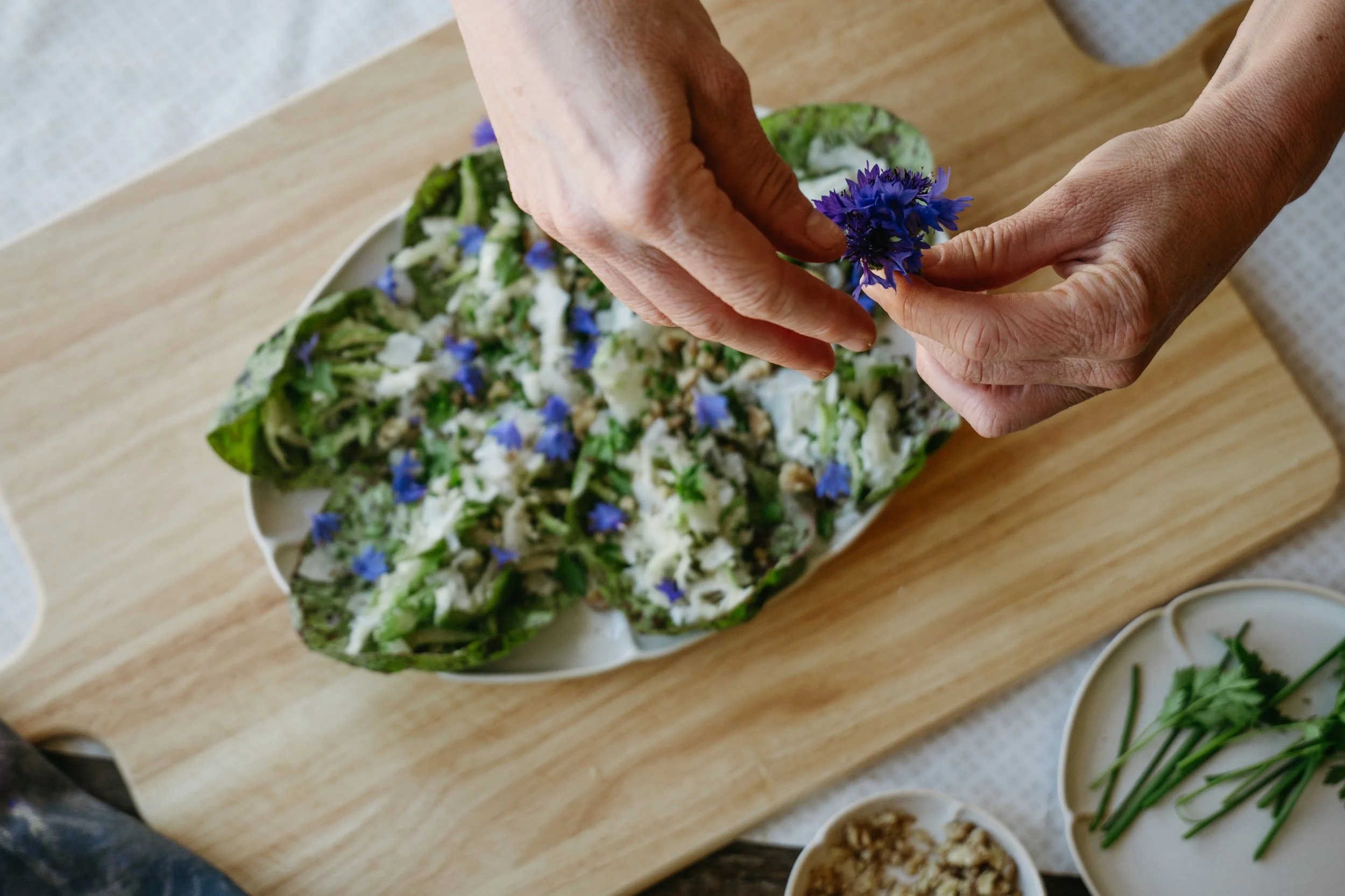 Person garnishing a salad with a purple flower on a wooden table. The salad contains greens and edible flowers. Several small bowls of salad toppings or ingredients are visible nearby.