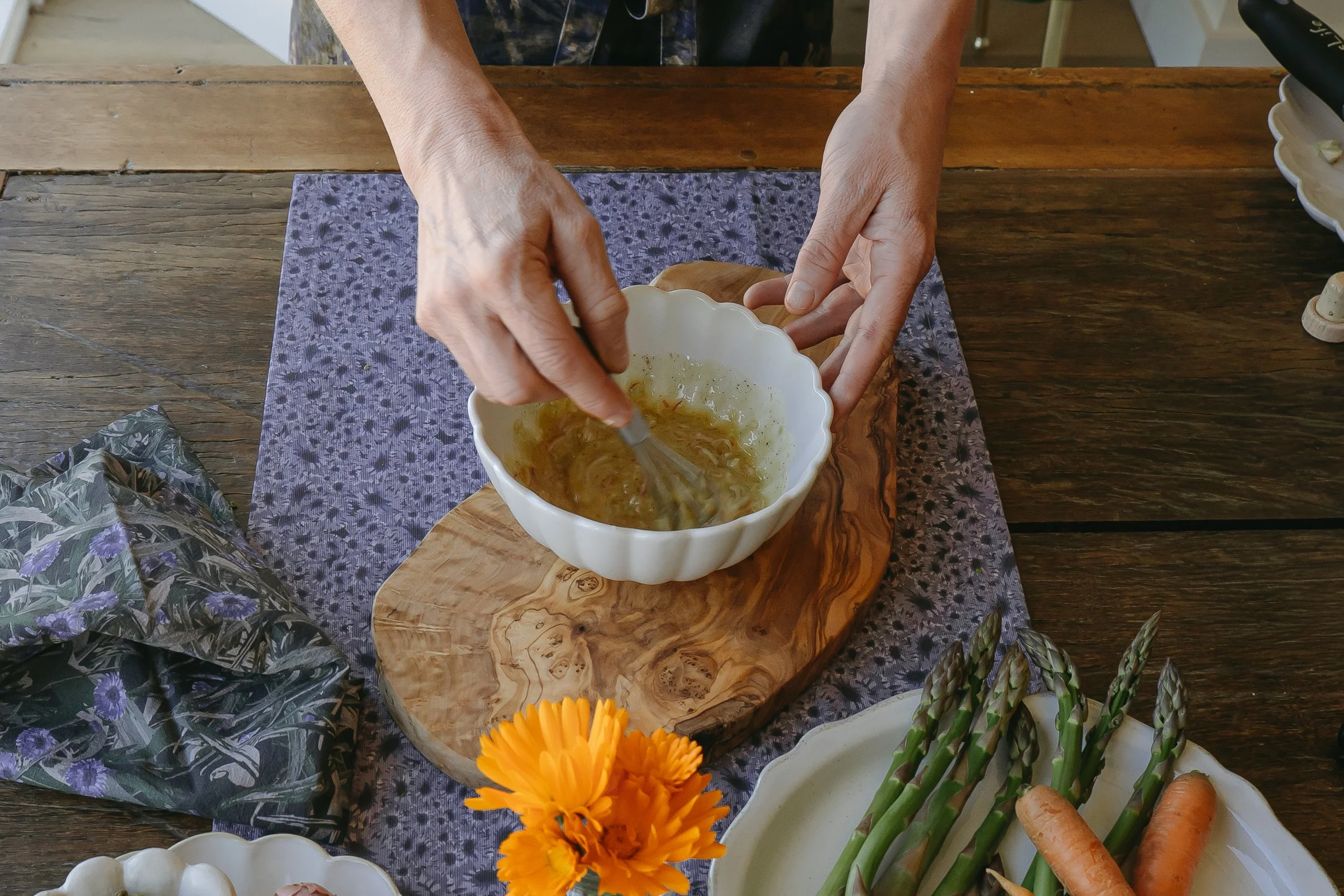 Hands whisking a liquid mixture in a white scalloped bowl on a wooden table, with a purple cloth, a pine cone, orange flowers, and vegetables nearby.