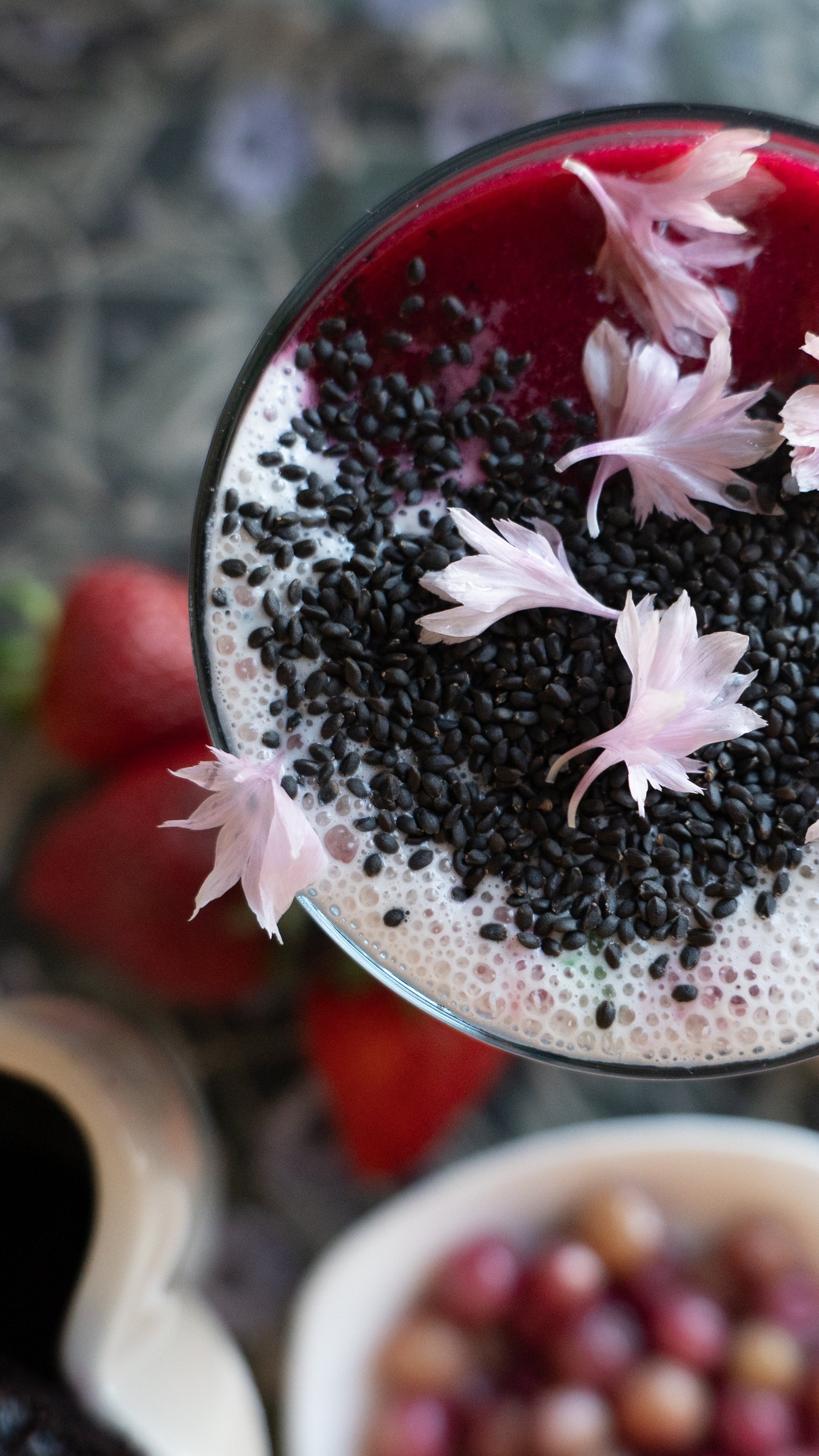 A top-down view of a glass containing a beverage topped with black sesame seeds and pink flower petals, with blurred bowls of small round snacks or berries in the background.