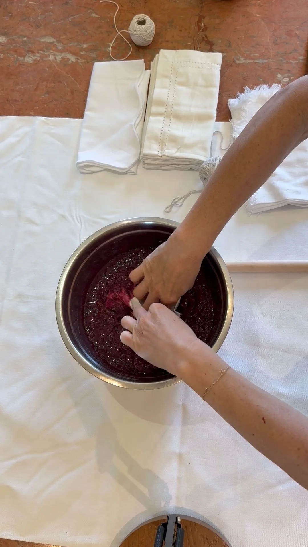 Two hands swirl a red dye in a metal bowl on a white tablecloth, with folded white fabric and a ball of string in the background.
