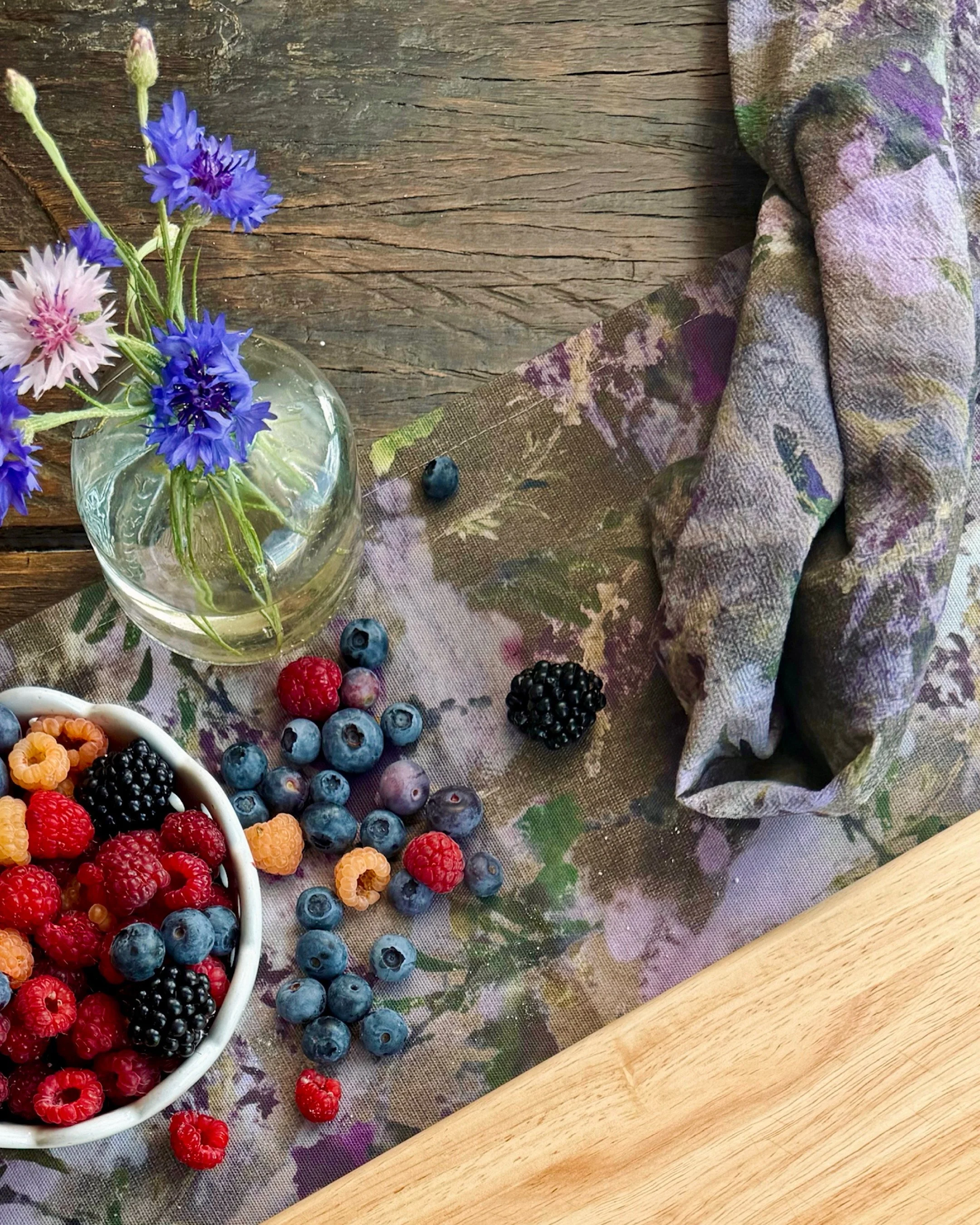 A wooden table with a signature Snacky Girl Snapdragon print, a small bouquet of blue and pink flowers in a glass vase, a bowl of mixed berries including blueberries, raspberries, blackberries, and golden raspberries, and a rolling pin.