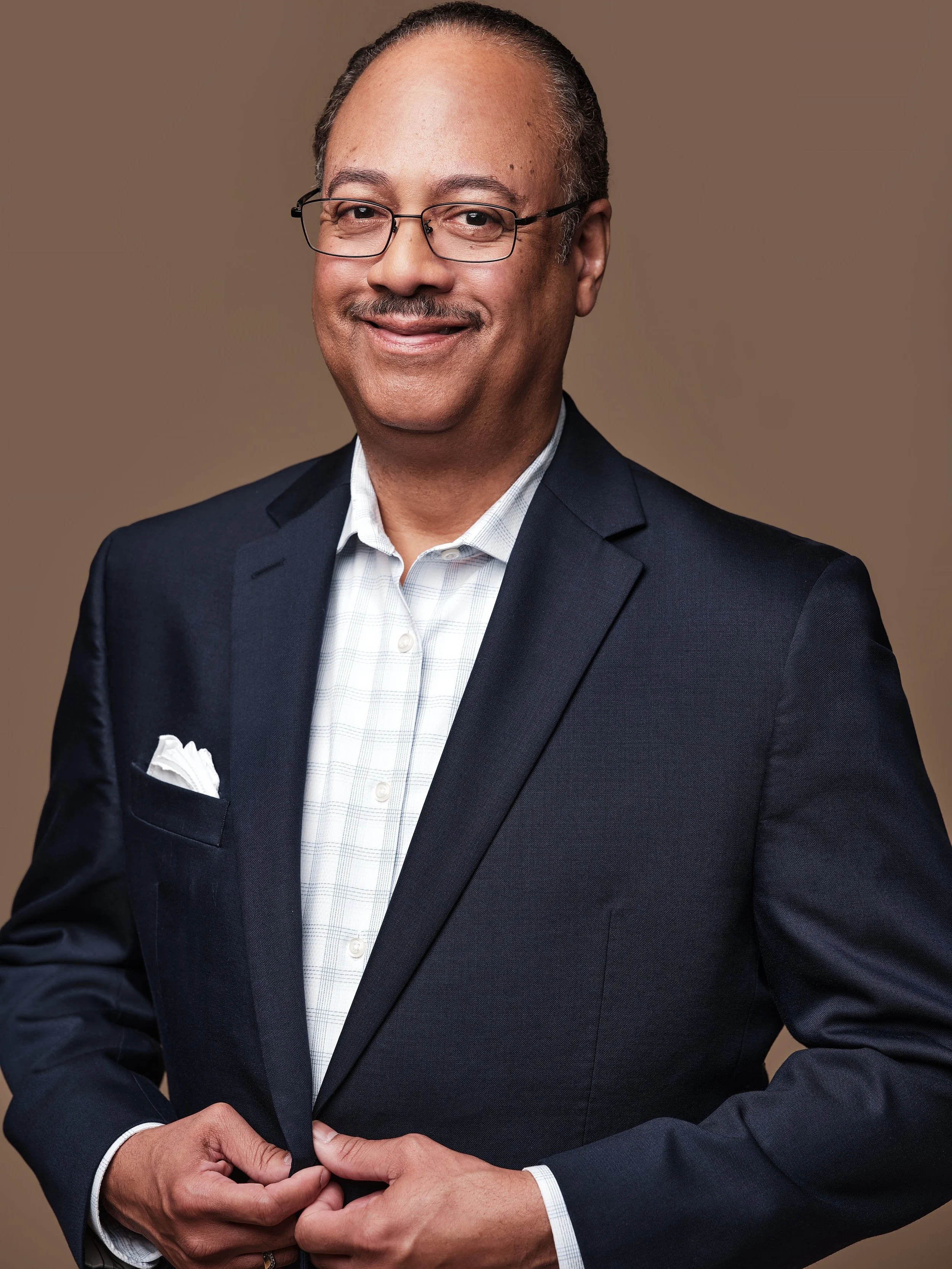 A smiling man wearing glasses, a navy blazer, white shirt, and white pocket square, standing against a beige background.