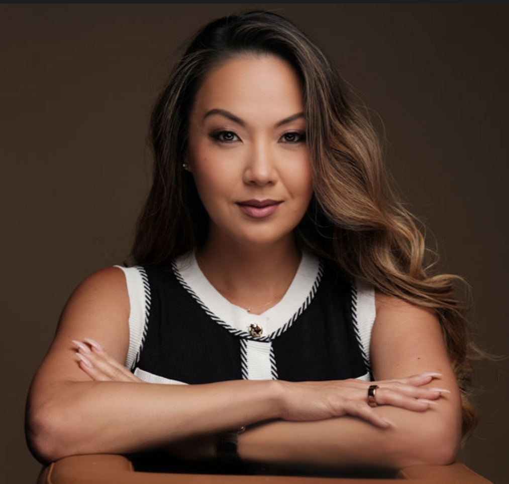 A woman with long light brown hair smiling, wearing a black top and blazer with gold buttons, accessorized with pearl necklaces, earrings, and a delicate necklace, posed with her hand on her hip against a plain background.