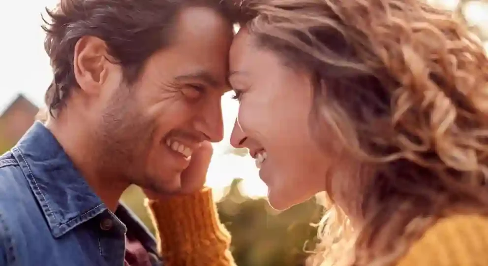 A man and woman with their foreheads touching, smiling and sharing a joyful moment outdoors during sunset or golden hour.