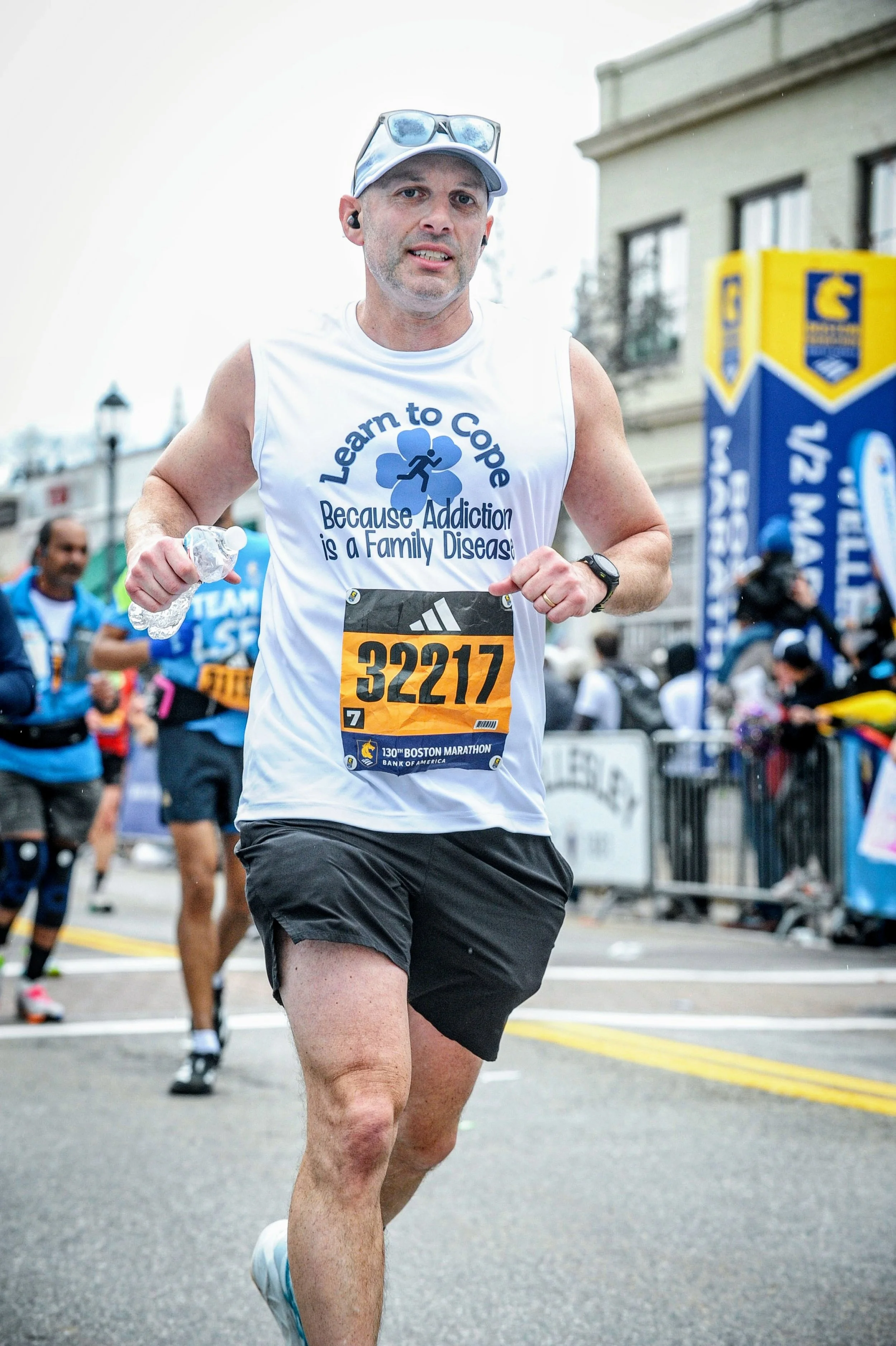 Runner wearing a recovery-themed shirt and Boston Marathon bib, holding a water bottle during a road race.