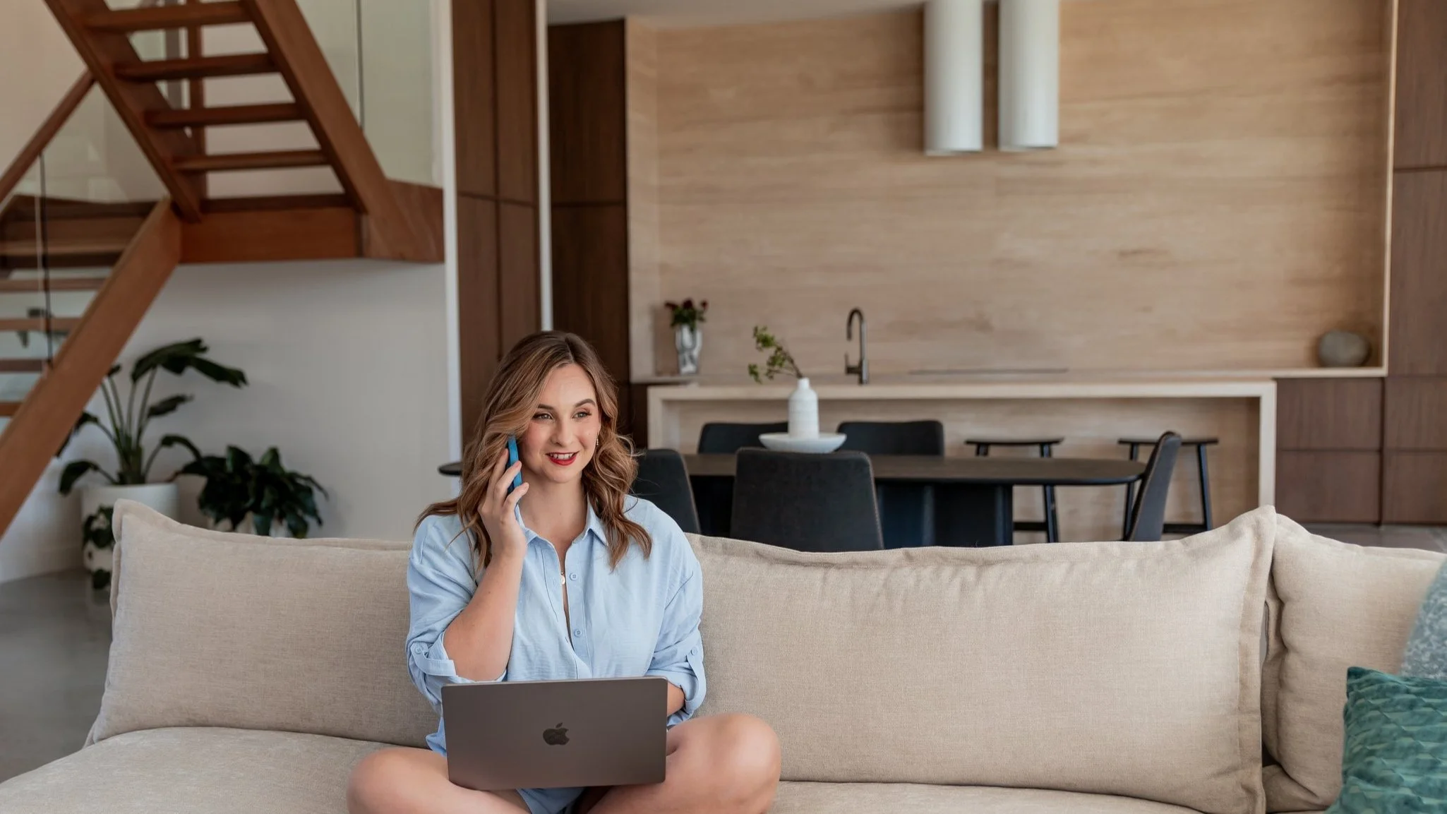 A woman sitting on a beige sofa in a modern living room, holding a phone to her ear and using a laptop on her lap, with a kitchen and dining area in the background.