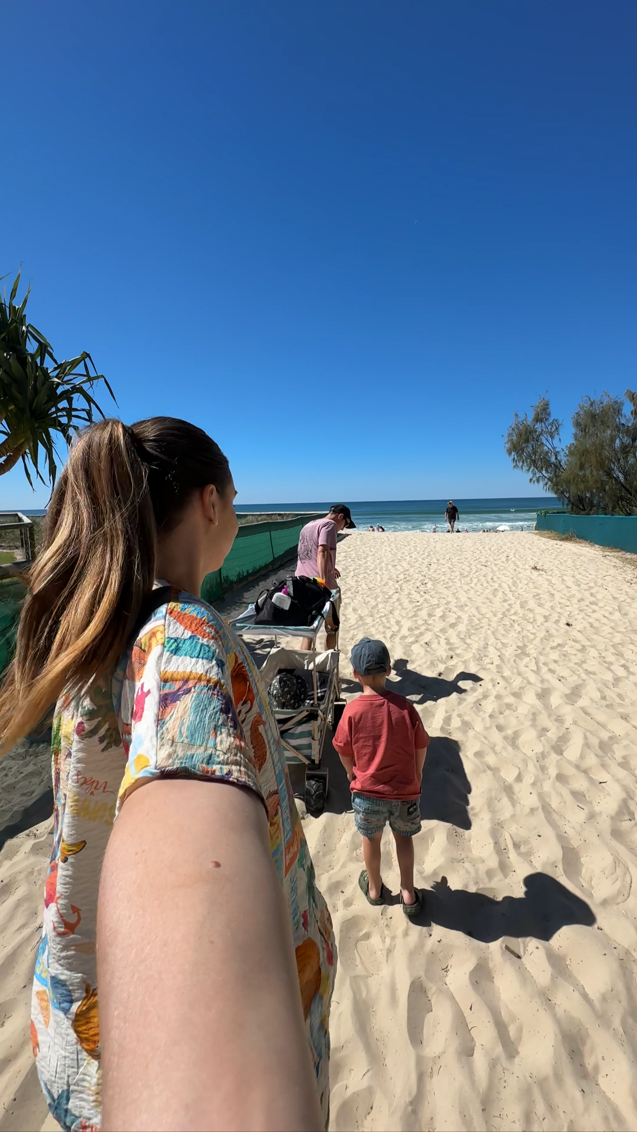 People walking on a sandy beach pathway toward the ocean, with some carrying belongings and children, under a clear blue sky.