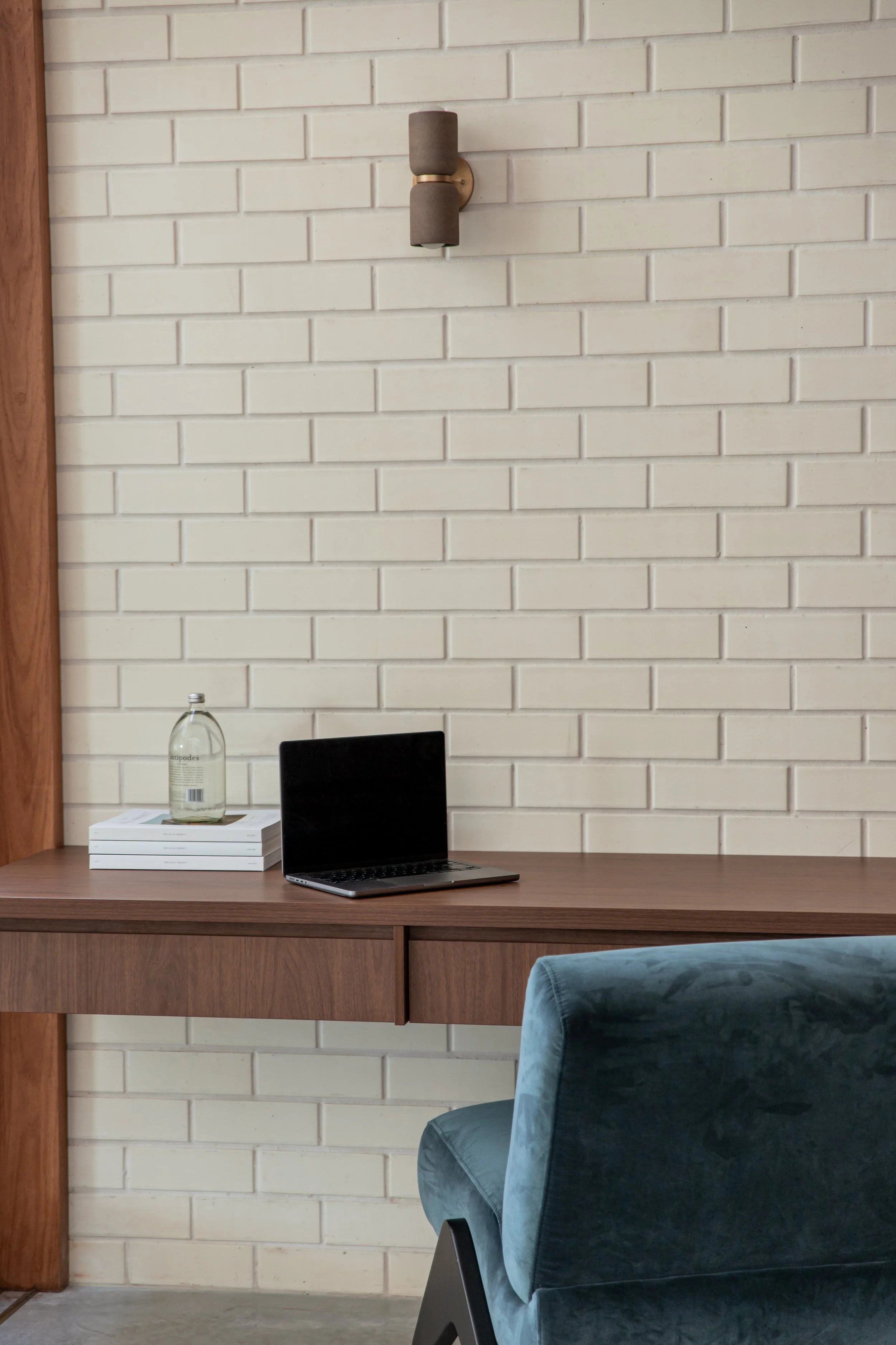 A wooden desk with a laptop, a clear glass water bottle, and three stacked books. A brick wall in the background and a blue velvet chair in the foreground.