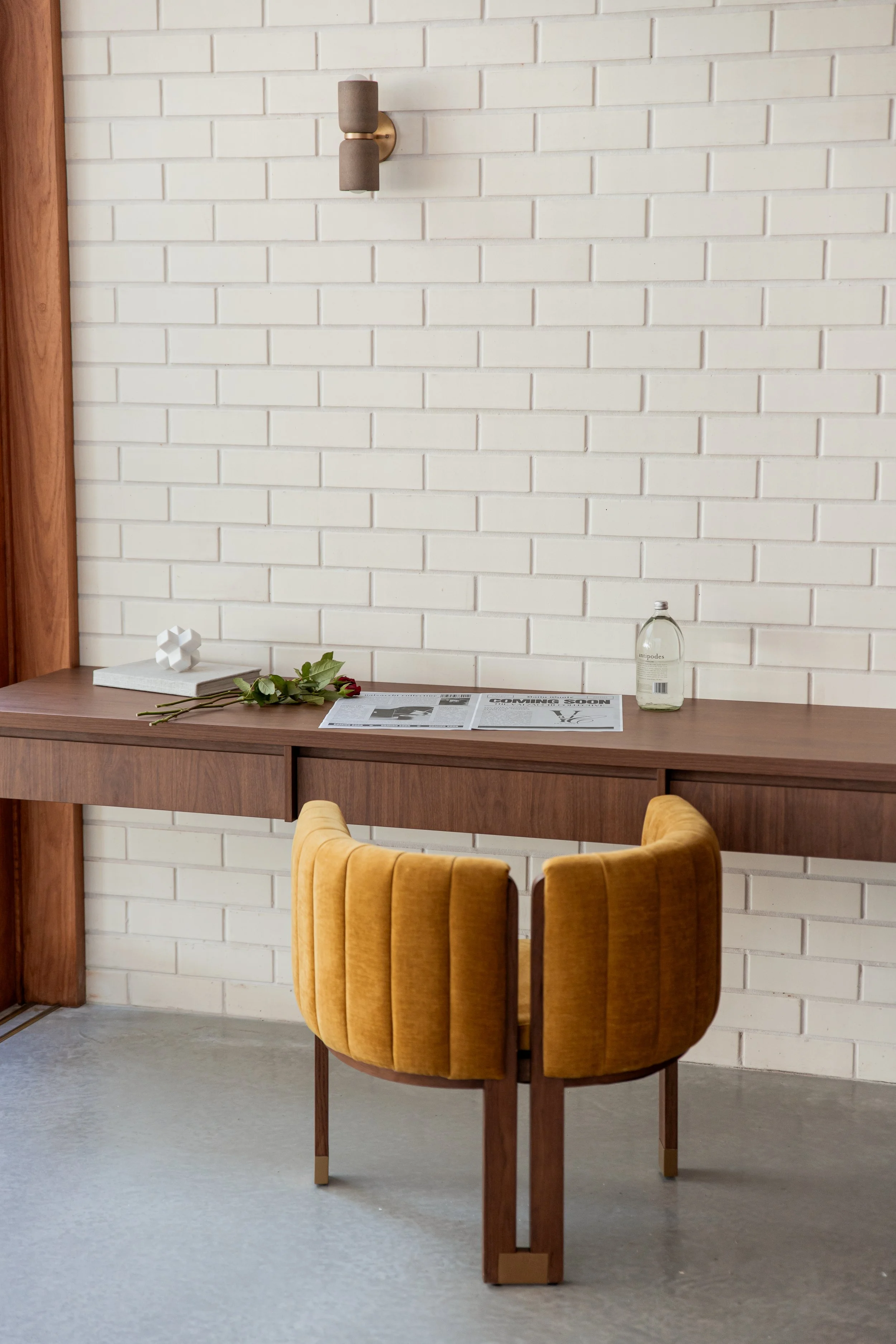A wooden table with a bouquet of flowers, a white decorative object, a bottle of water, and a newspaper on it, against a white brick wall with wall-mounted lighting and a mustard yellow upholstered chair in the foreground.