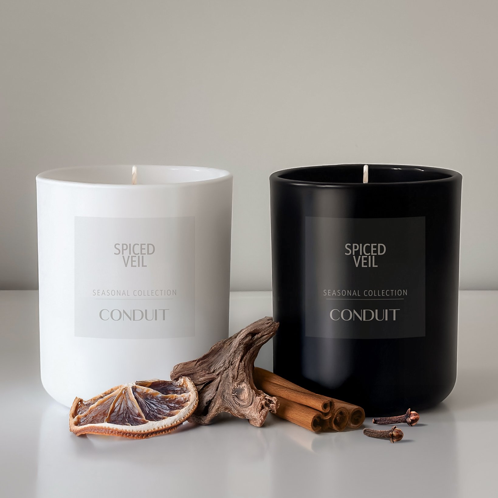 Two candles labeled 'Spiced Veil' from Seasonal Collection, one white and one black, placed on a white surface with dried orange slice, cinnamon sticks, a piece of driftwood, and cloves in front of them.